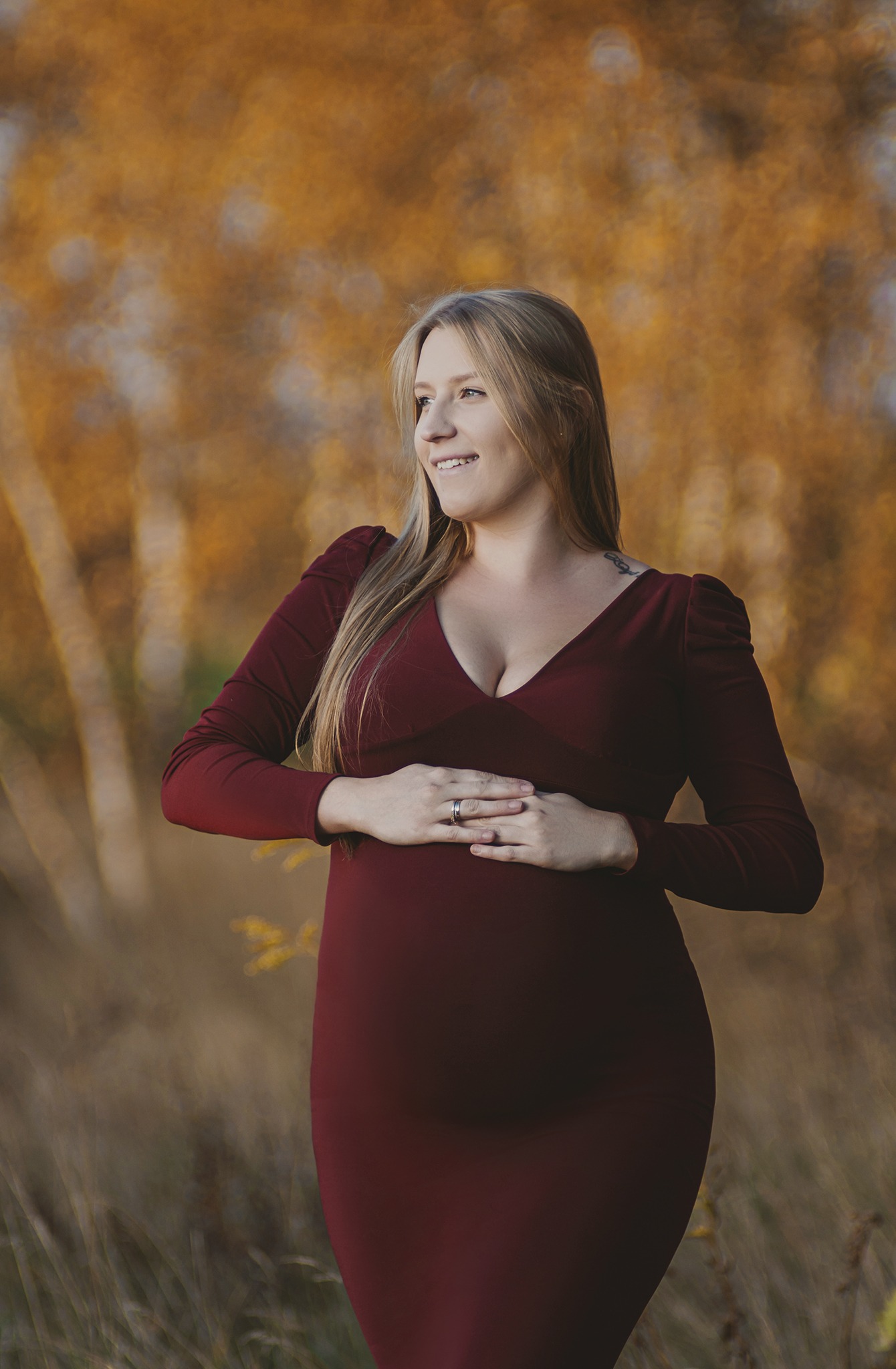 Kobieta w bordowej sukience ciążowej, trzymająca ręce na brzuchu, uśmiechnięta, z widocznym tatuażem na ramieniu, na tle jesiennych drzew.