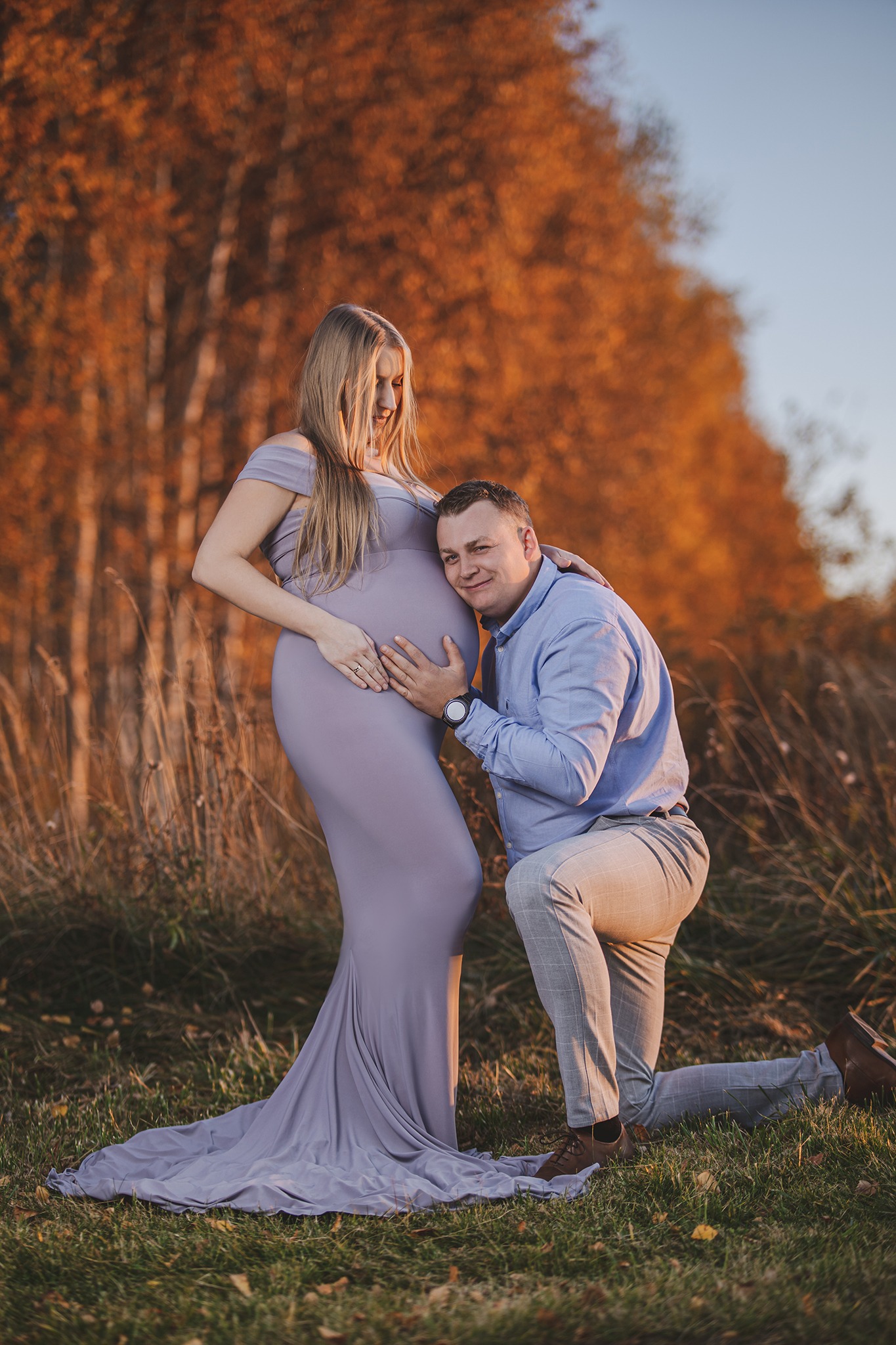 Sesja ciążowa w plenerze: przyszła mama w długiej, liliowej sukni i przyszły tata klęczący obok, przykładający ucho do brzucha, jesienne tło z drzewami o pomarańczowych liściach.