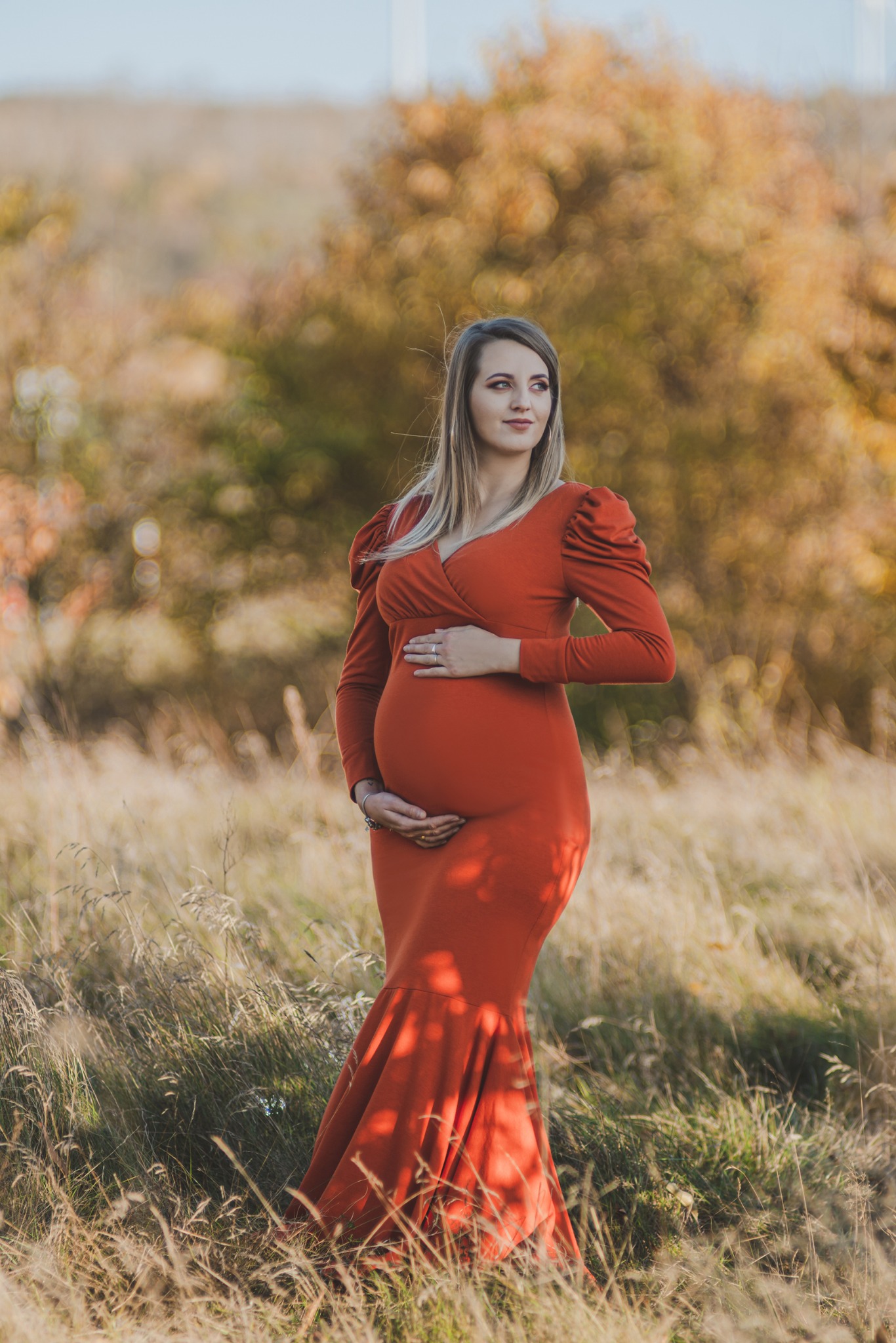 Kobieta w pomarańczowej sukni ciążowej, pozująca na tle jesiennej łąki z suchą trawą i drzewami w tle.