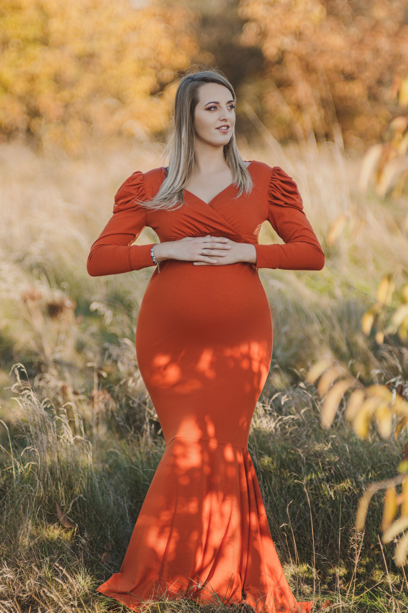 Kobieta w dopasowanej, pomarańczowej sukni z rękawami typu bufki, delikatnie trzymająca dłonie na brzuchu, pozująca na tle łąki z jesiennymi drzewami w tle.