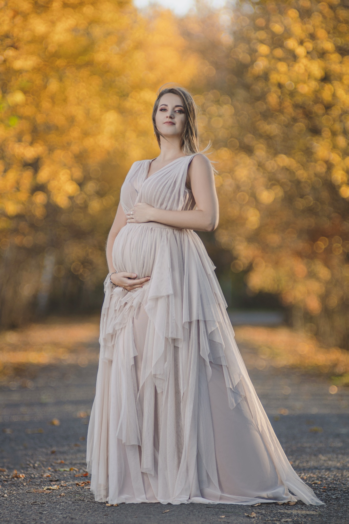 Kobieta w ciąży w długiej, zwiewnej sukni pozuje na tle jesiennych drzew z rozproszonym światłem.
