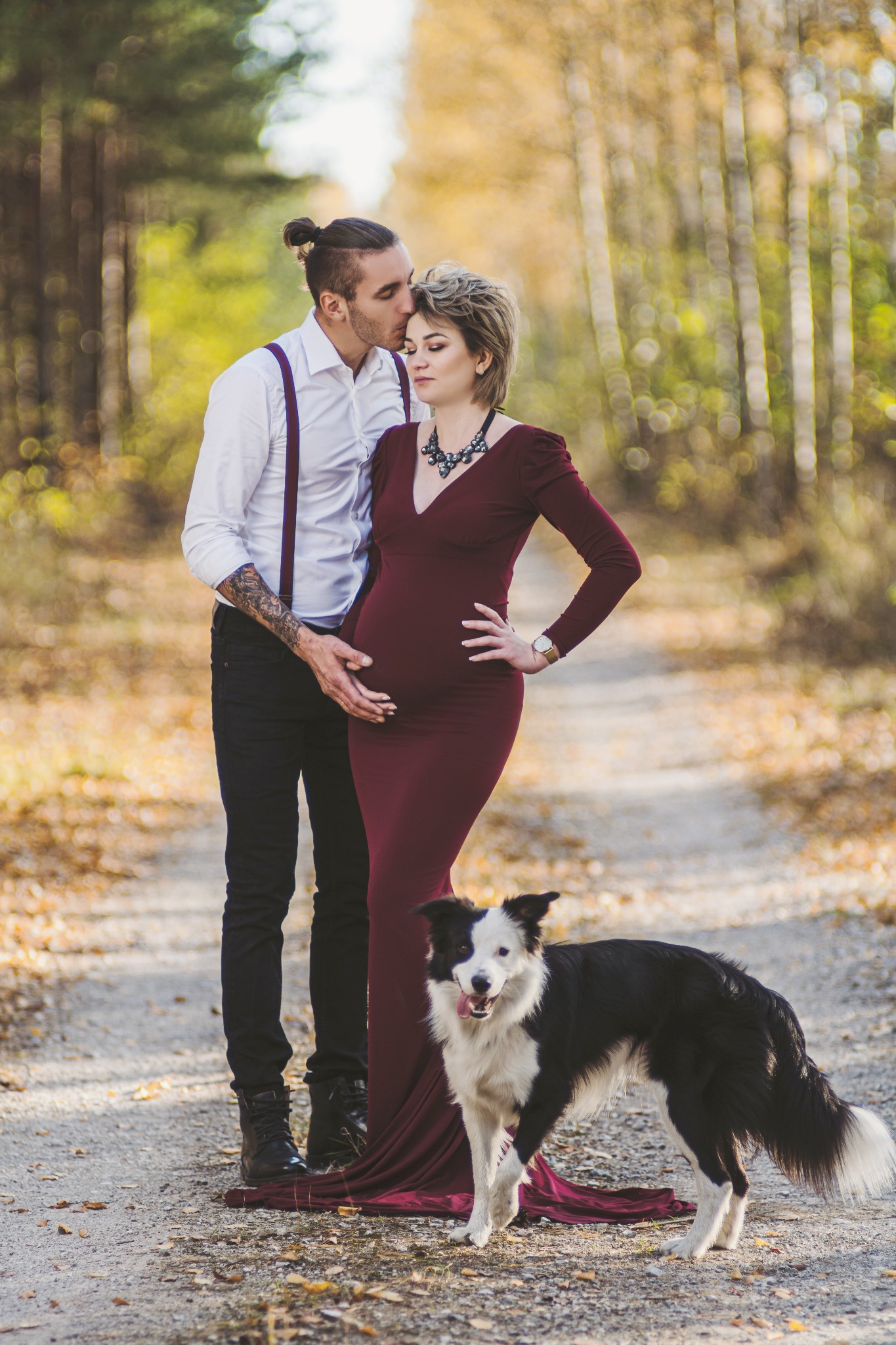 Mężczyzna z tatuażem na ramieniu całuje w czoło kobietę w bordowej sukni ciążowej, obok nich pies rasy border collie na leśnej ścieżce.
