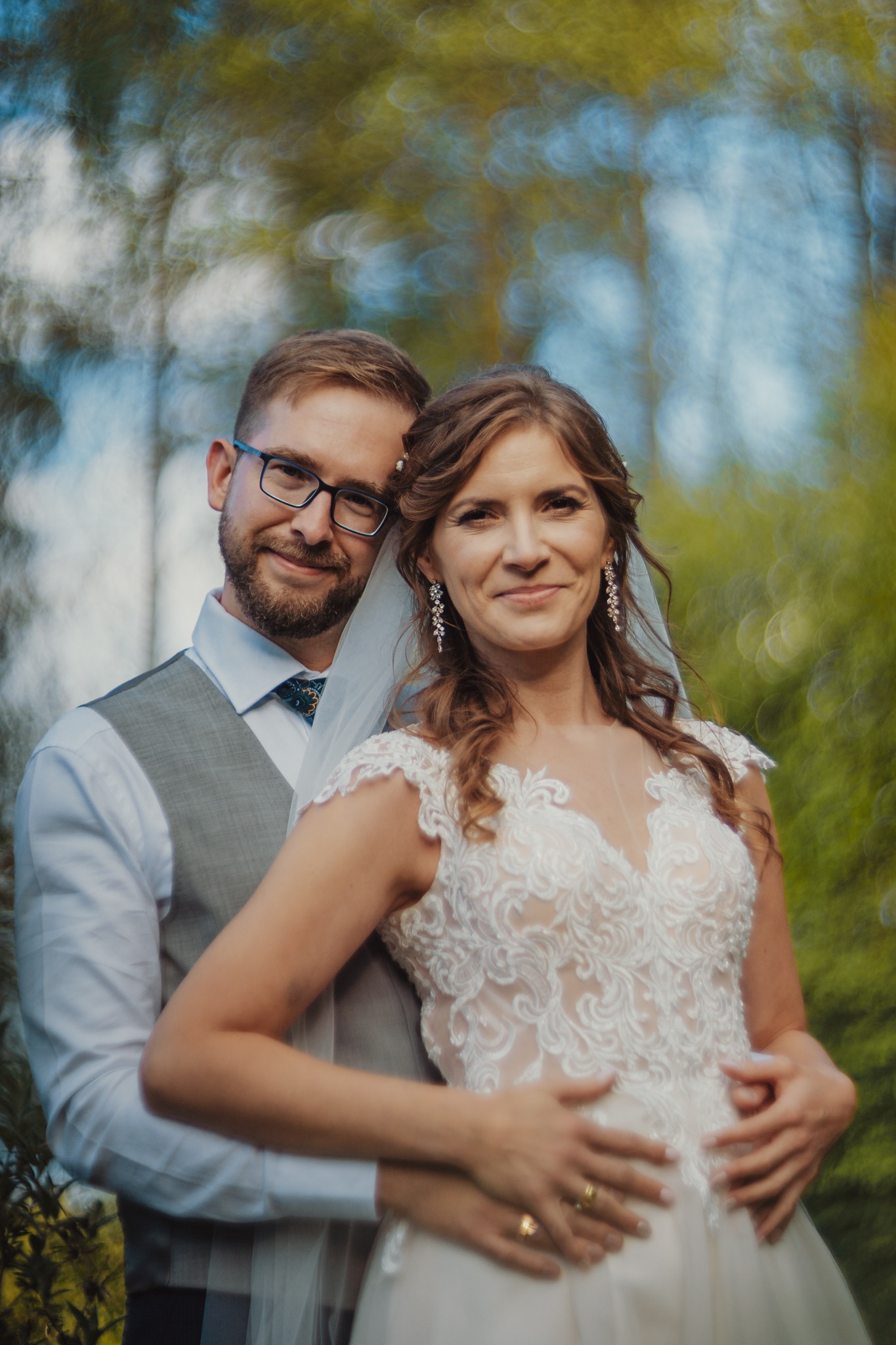 Uśmiechnięta para młoda w objęciach na tle rozmytej zieleni, panna młoda w koronkowej sukni z dekoltem w serek i długimi kolczykami, pan młody w szarej kamizelce i okularach.