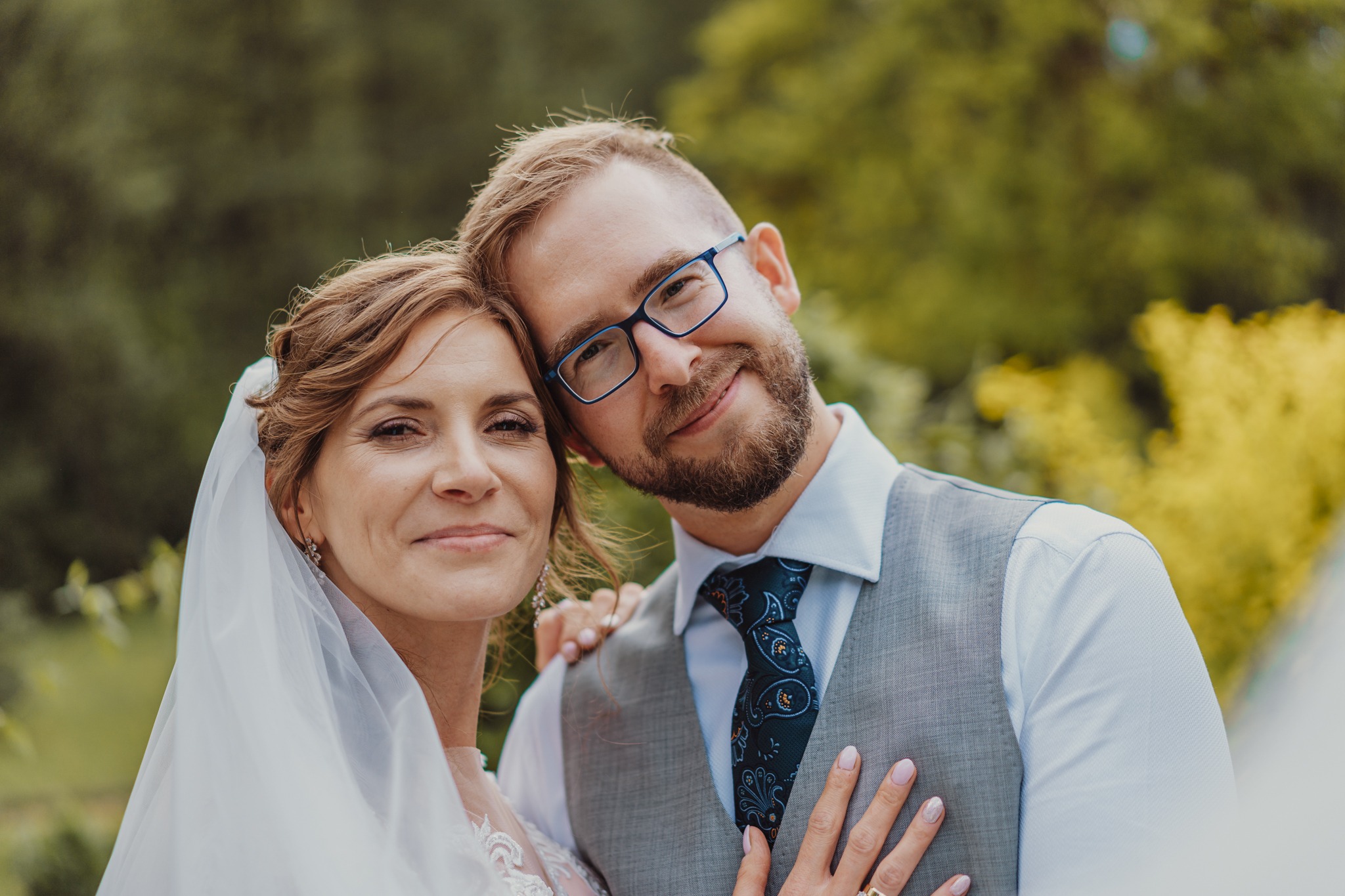 Uśmiechnięta para młoda w zbliżeniu: panna młoda w welonie i sukni z koronką, pan młody w okularach, kamizelce i wzorzystym krawacie, zielone tło.