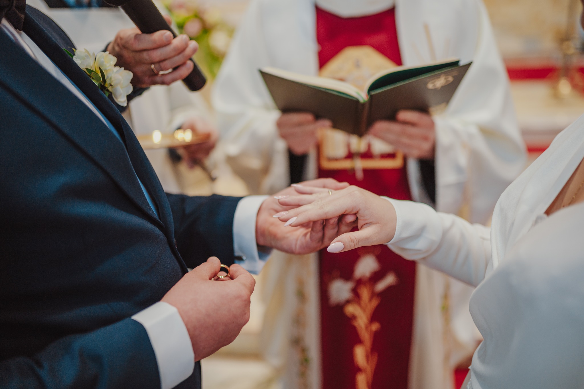 Moment zakładania obrączki podczas ceremonii ślubnej w kościele w Węgrzcach Wielkich, widoczny fragment garnituru pana młodego z butonierką oraz dłoń panny młodej z pierścionkiem zaręczynowym.
