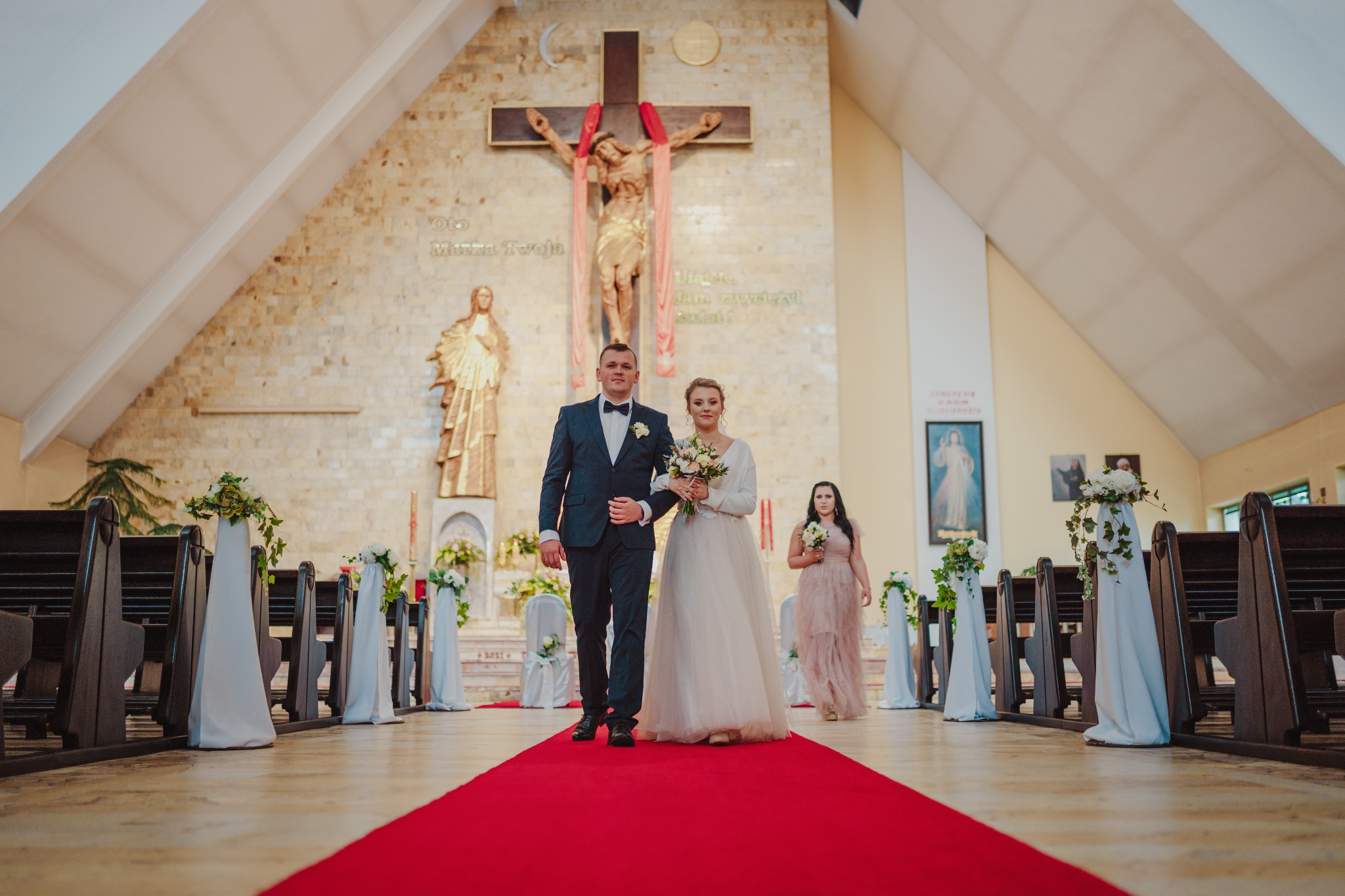 Para młoda idąca przez kościół po czerwonym dywanie, dekoracje kwiatowe na ławkach, w tle krucyfiks i postać kobiety w sukni.