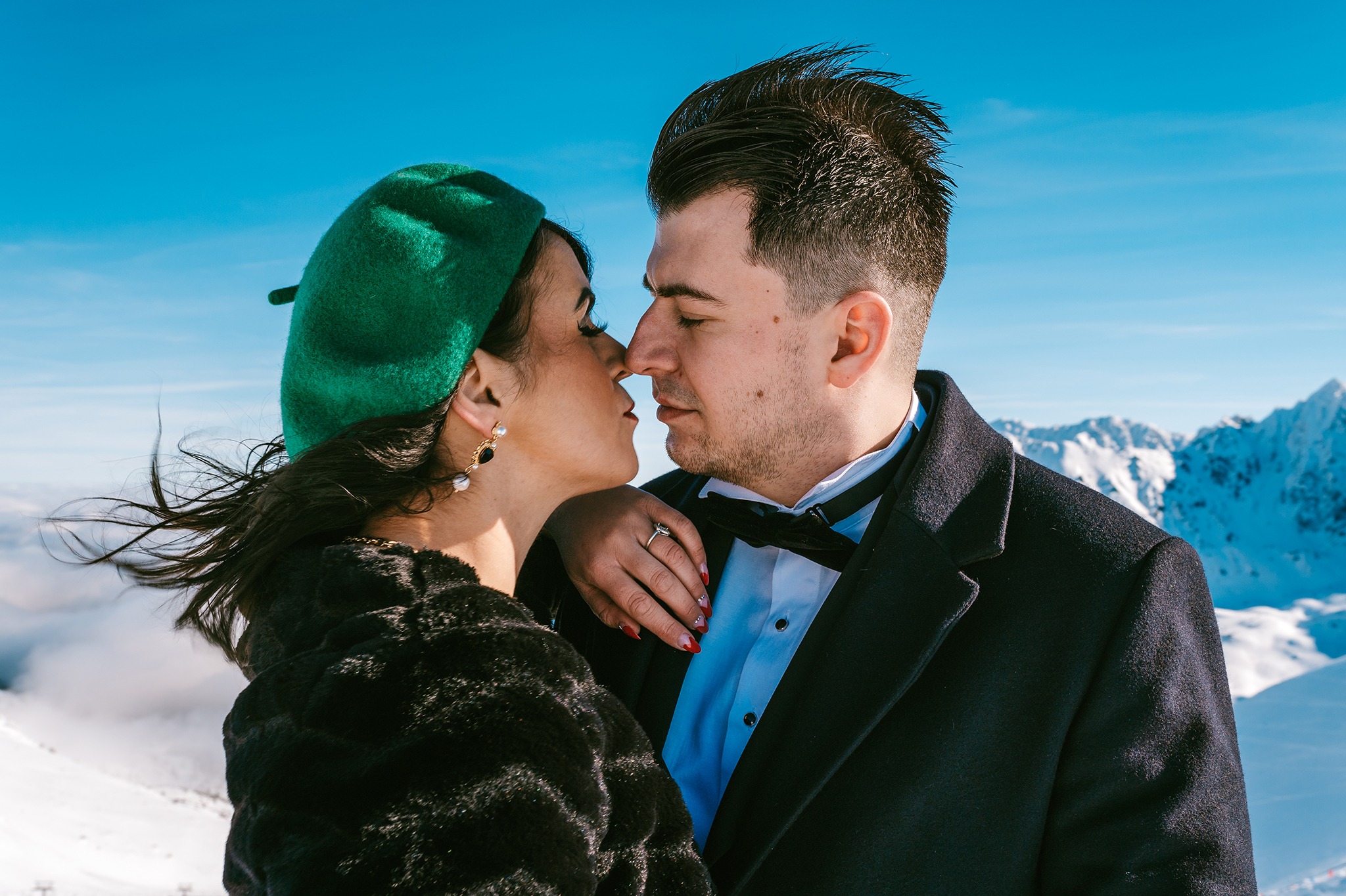 Elegancka para w strojach ślubnych, delikatnie dotykająca się nosami na tle ośnieżonych gór i błękitnego nieba. Kobieta w zielonym berecie i futrze, mężczyzna w smokingu z muszką.