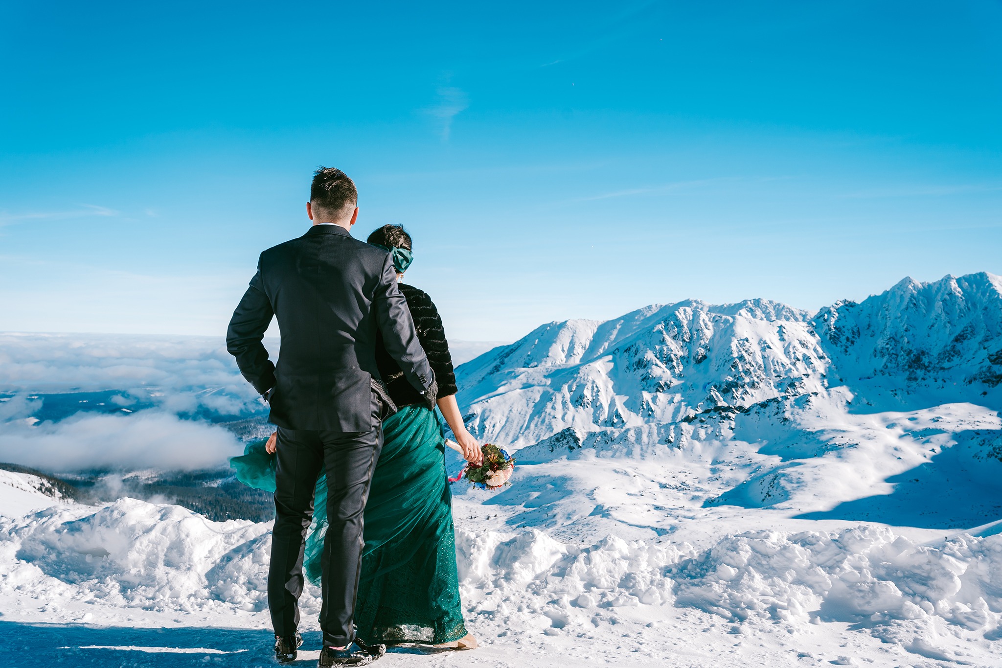 Para młoda w strojach ślubnych, stojąca na śniegu z widokiem na ośnieżone góry w słoneczny dzień. Pan młody w ciemnym garniturze, panna młoda w szmaragdowej sukni trzyma bukiet.