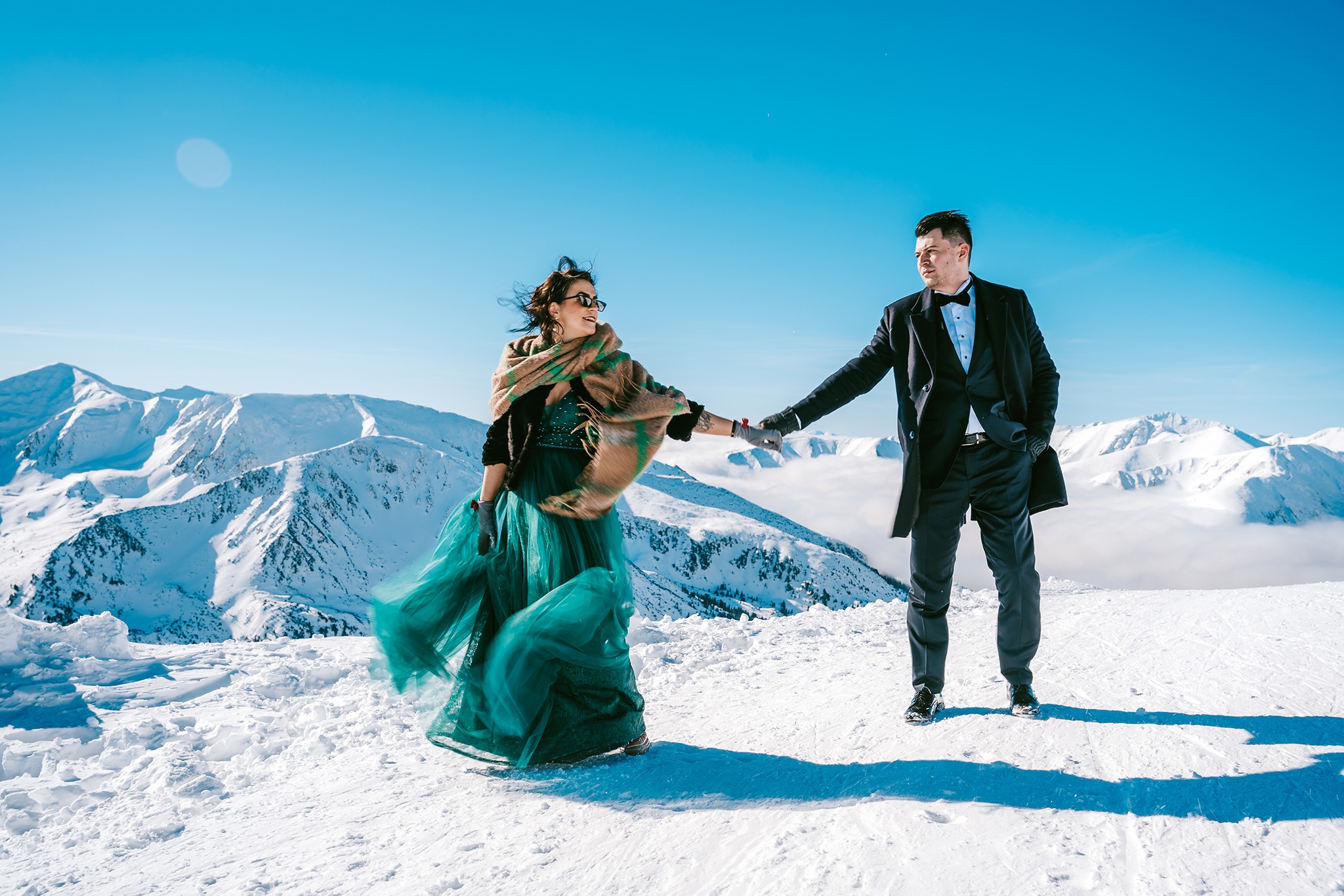 Elegancko ubrana para trzymająca się za ręce na tle ośnieżonych gór i błękitnego nieba, kobieta w turkusowej sukni i szalu, mężczyzna w czarnym garniturze z muszką.