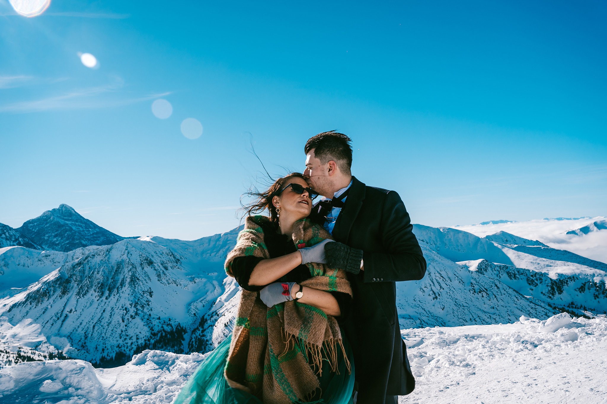 Para młoda w zimowej scenerii górskiej: panna młoda w okularach przeciwsłonecznych i zielonej sukni, owinięta w pled, pan młody w smokingu całuje ją w czoło na tle ośnieżonych szczytów i błękitnego...