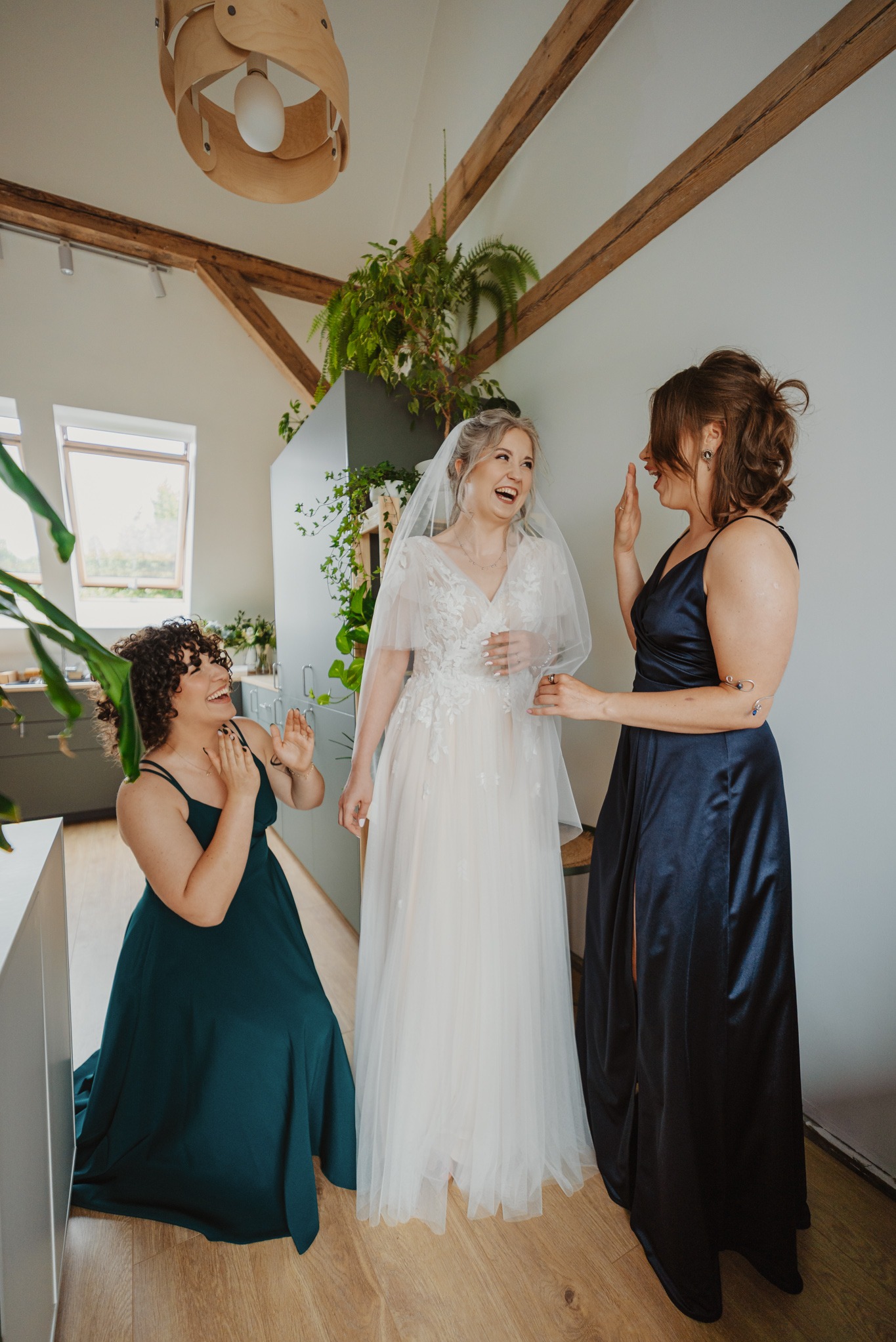 Uśmiechnięta panna młoda w koronkowej sukni i welonie, otoczona dwiema druhnami w długich sukniach, w jasnym wnętrzu z drewnianymi belkami i roślinami.