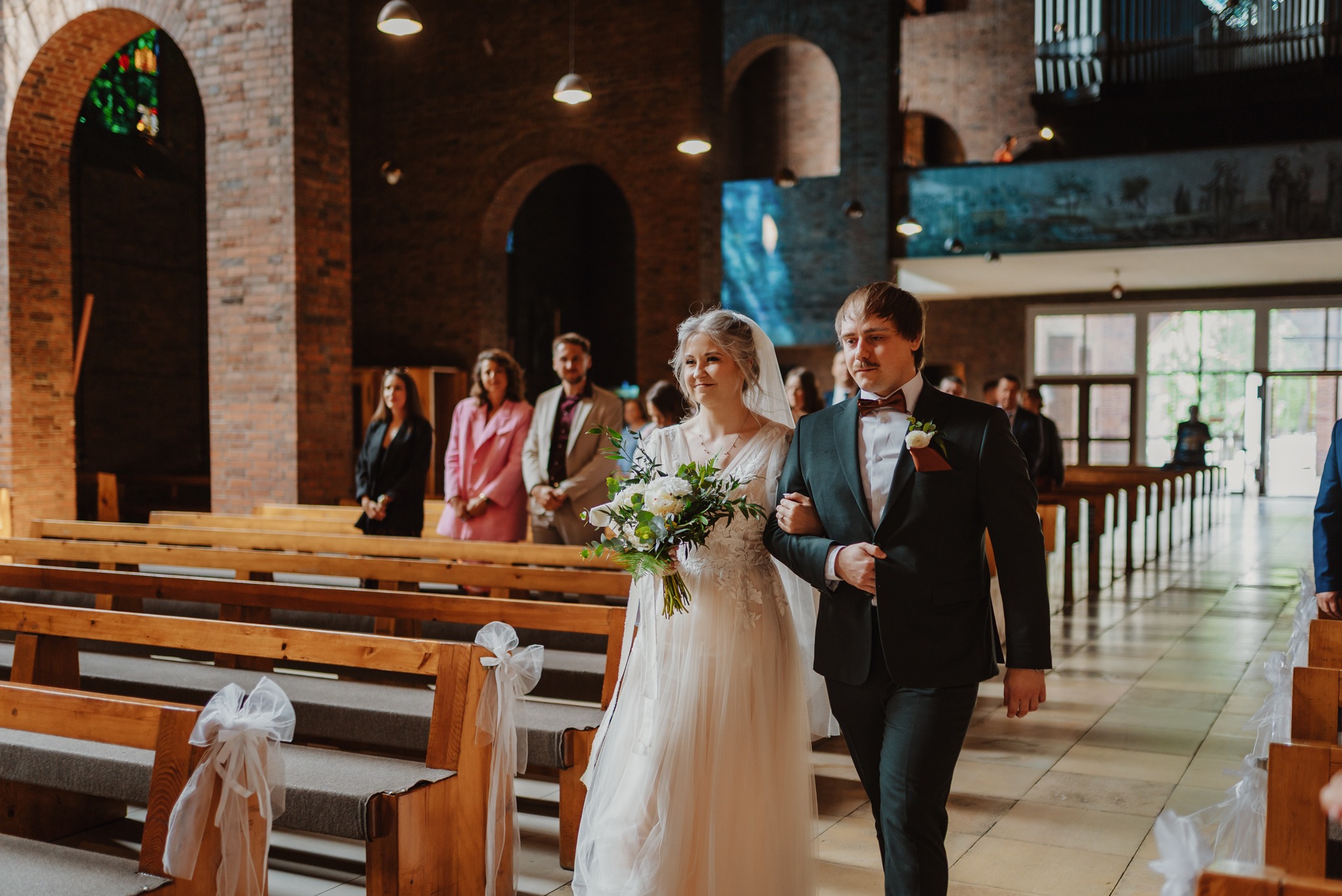 Para młoda w kościele, idąca wzdłuż ławek udekorowanych białymi kokardami. Panna młoda trzyma bukiet, a pan młody ma bordową muchę. W tle goście.