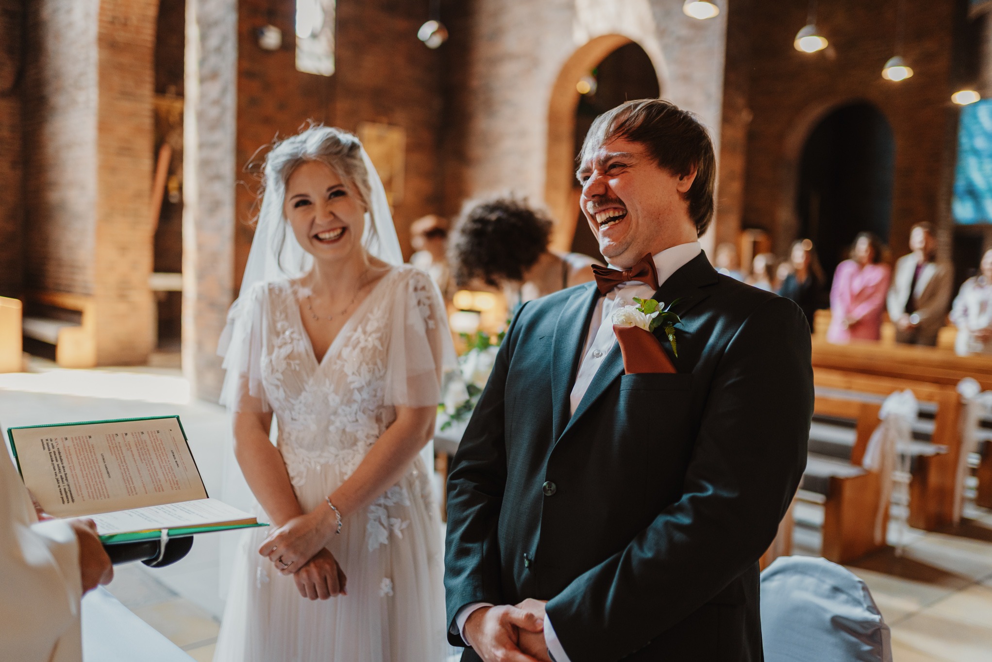 Uśmiechnięta para młoda podczas ceremonii ślubnej w kościele. Panna młoda w koronkowej sukni, pan młody w ciemnym garniturze z muszką. W tle goście i elementy architektury sakralnej.
