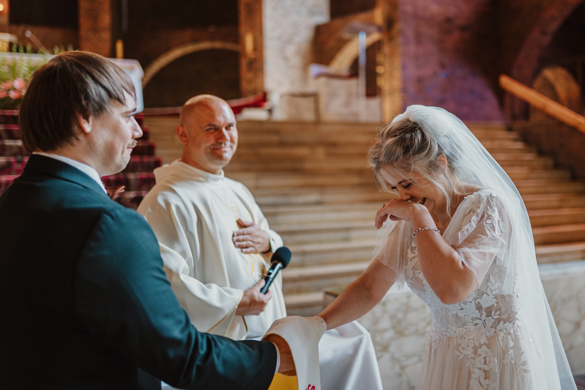 Uśmiechnięta panna młoda w koronkowej sukni podczas ceremonii ślubnej w kościele, z księdzem i panem młodym w tle. Jasne, naturalne światło.