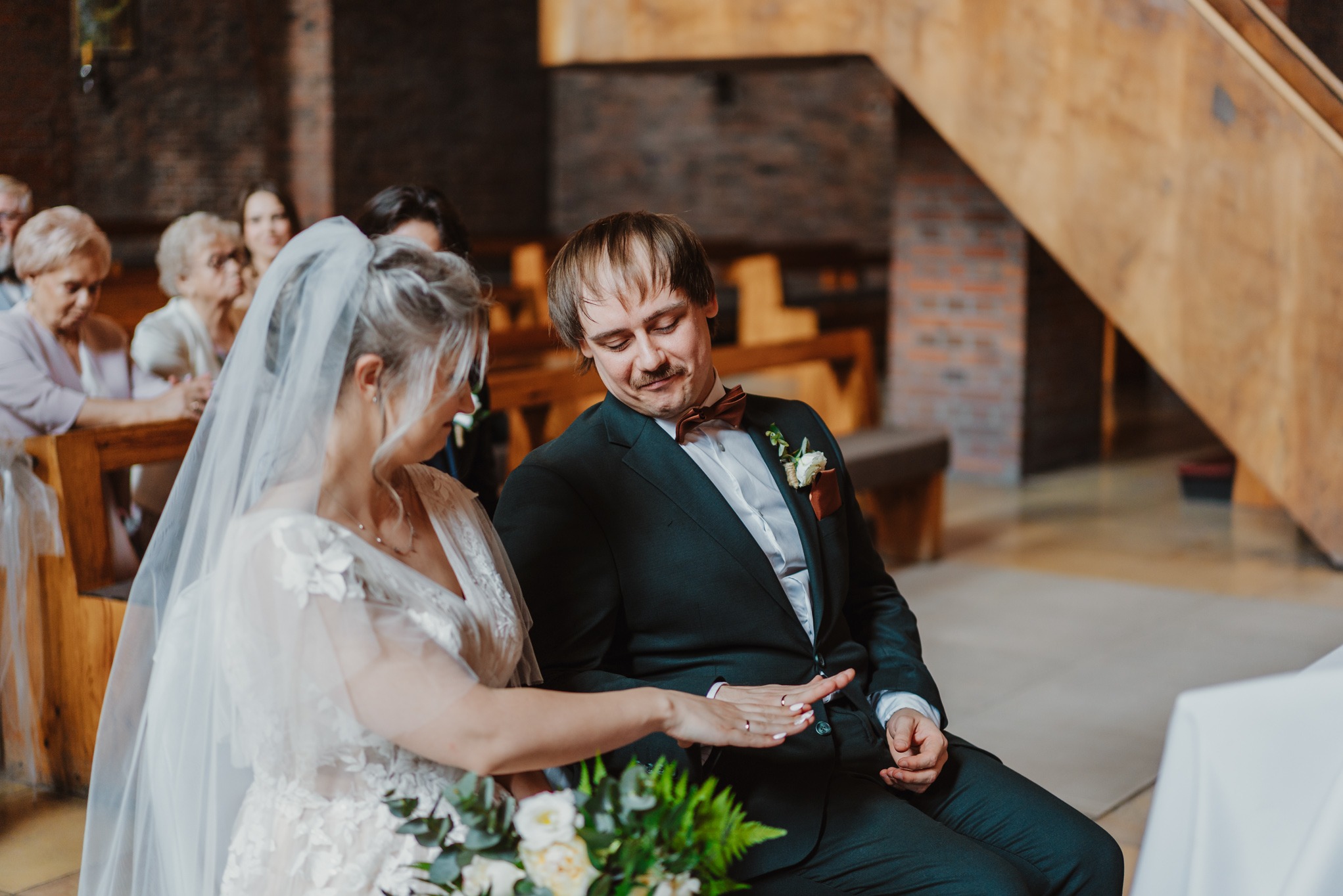 Para młoda siedzi obok siebie podczas ceremonii ślubnej w ceglanym wnętrzu. Panna młoda z welonem i bukietem, pan młody w ciemnym garniturze z muszką.