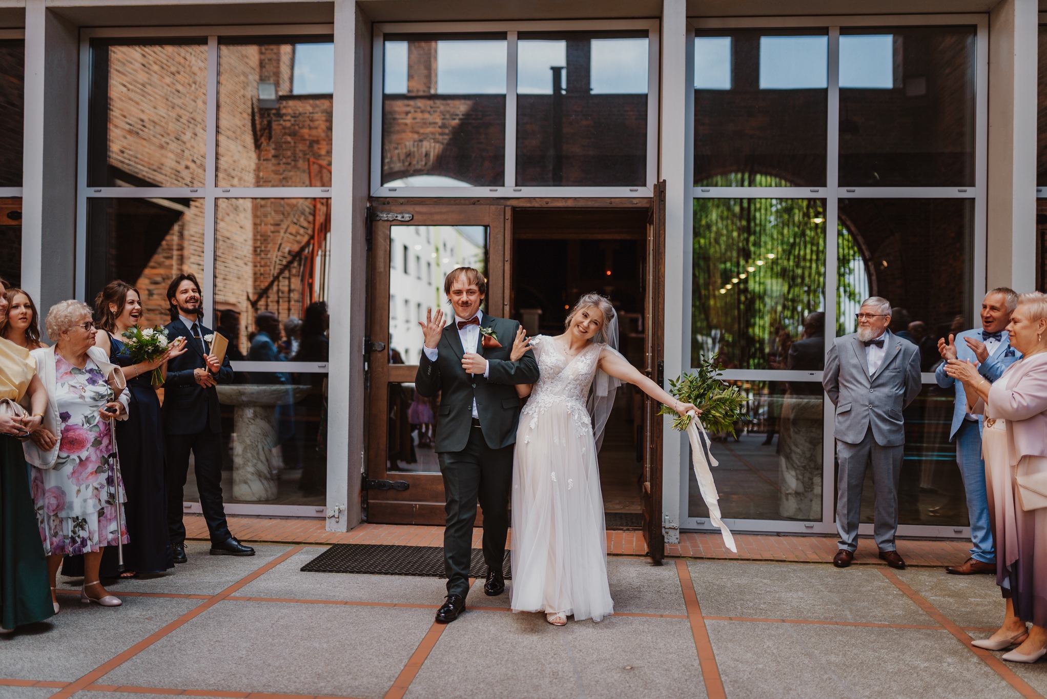 Uśmiechnięta para młoda opuszcza kościół, witana przez gości. Pan młody w ciemnym garniturze, panna młoda w białej sukni z bukietem. Architektura kościoła z cegły w tle.