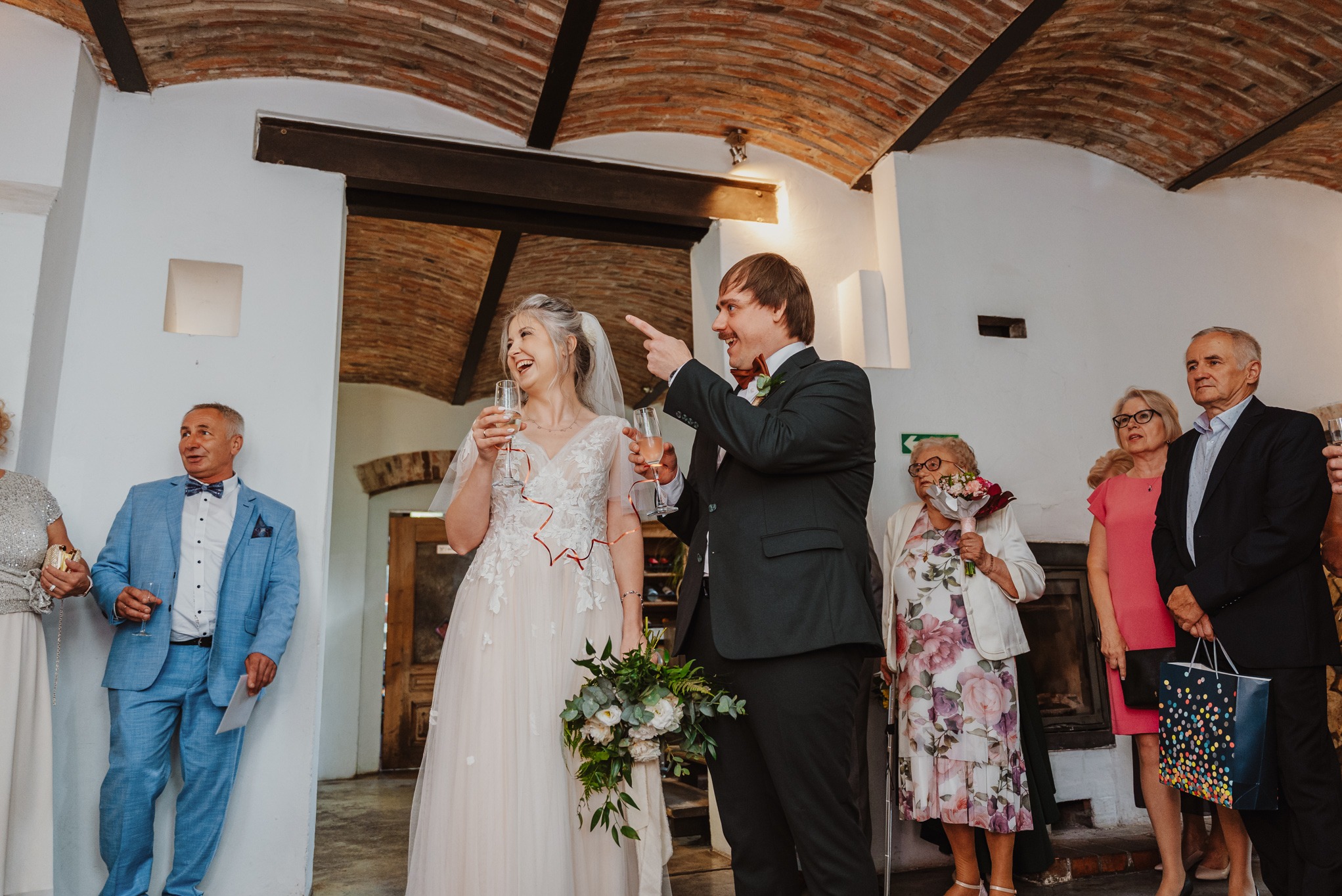 Uśmiechnięta para młoda w trakcie toastu na sali weselnej z ceglanym sklepieniem, w otoczeniu gości. Pan młody wskazuje palcem w górę, trzymając kieliszek szampana.
