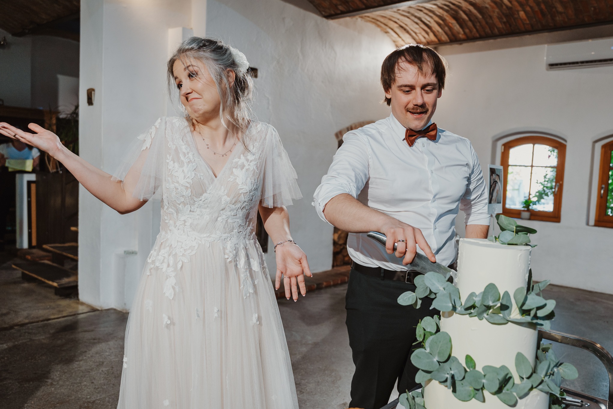 Uśmiechnięta para młoda kroi tort weselny w rustykalnej sali. Panna młoda w koronkowej sukni z uniesionymi rękami, pan młody w muszce z nożem nad tortem.