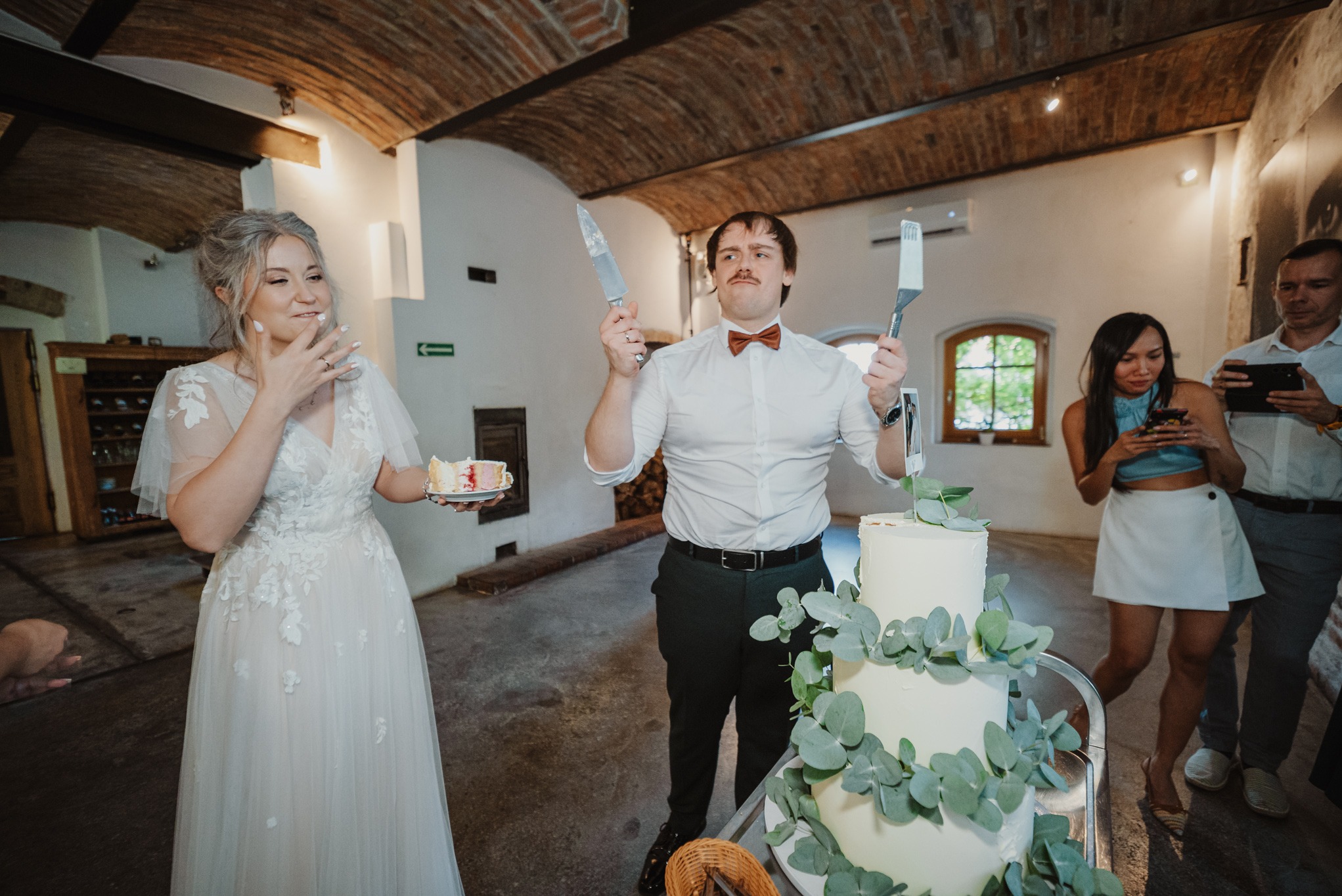 Uśmiechnięta para młoda kroi tort weselny. Pan młody z muszką trzyma nóż i łopatkę. Goście weselni obserwują i robią zdjęcia. Ceglane sklepienie w tle.