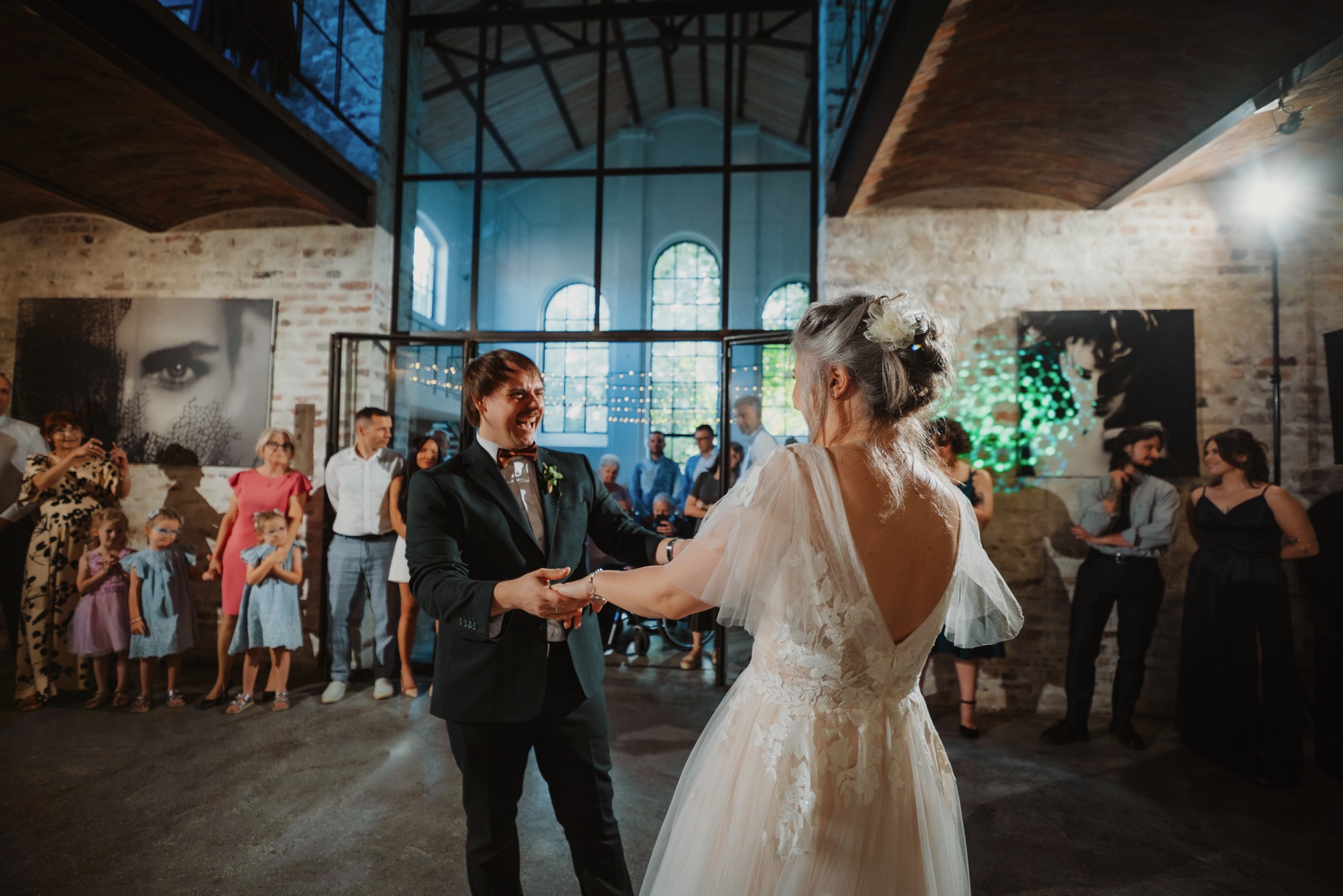 Uśmiechnięta para młoda w tańcu, otoczona gośćmi weselnymi w przestronnym wnętrzu z ceglanymi ścianami i dużymi oknami. Pan młody w ciemnym garniturze, panna młoda w sukni z odkrytymi plecami.