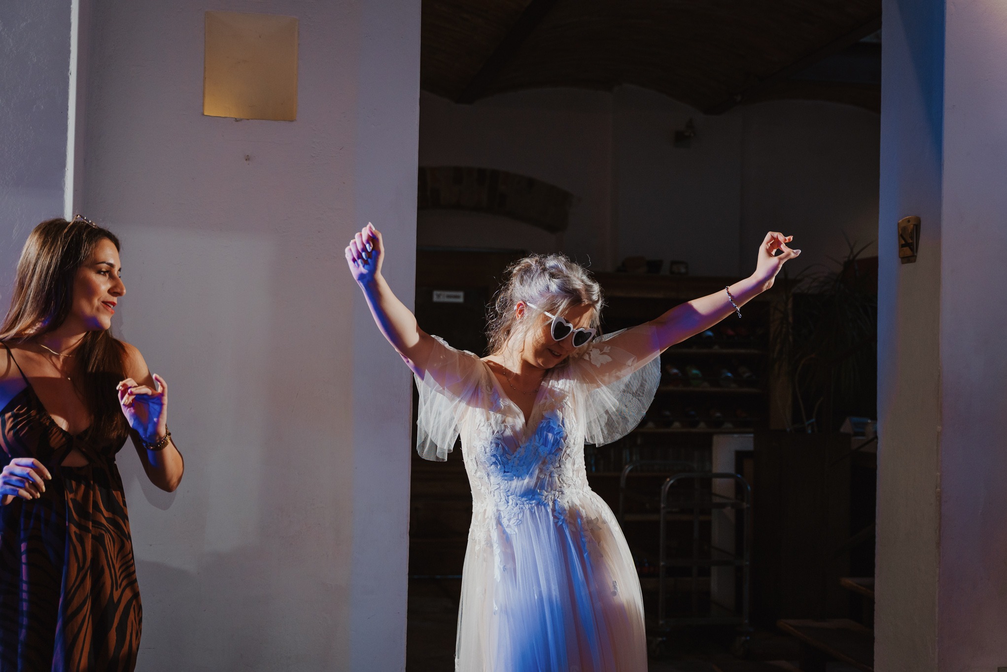 Rozbawiona panna młoda w okularach w kształcie serc tańczy z uniesionymi rękami, obok stoi uśmiechnięta kobieta w sukience w zebrę, w tle wnętrze sali weselnej.