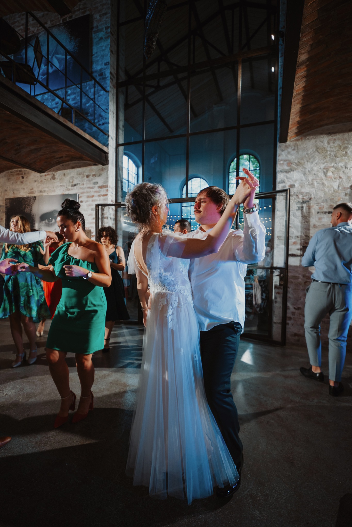 Para młoda w tańcu podczas wesela w industrialnym wnętrzu z gośćmi. Panna młoda w zwiewnej sukni, pan młody w białej koszuli. W tle ceglane ściany i duże okna.