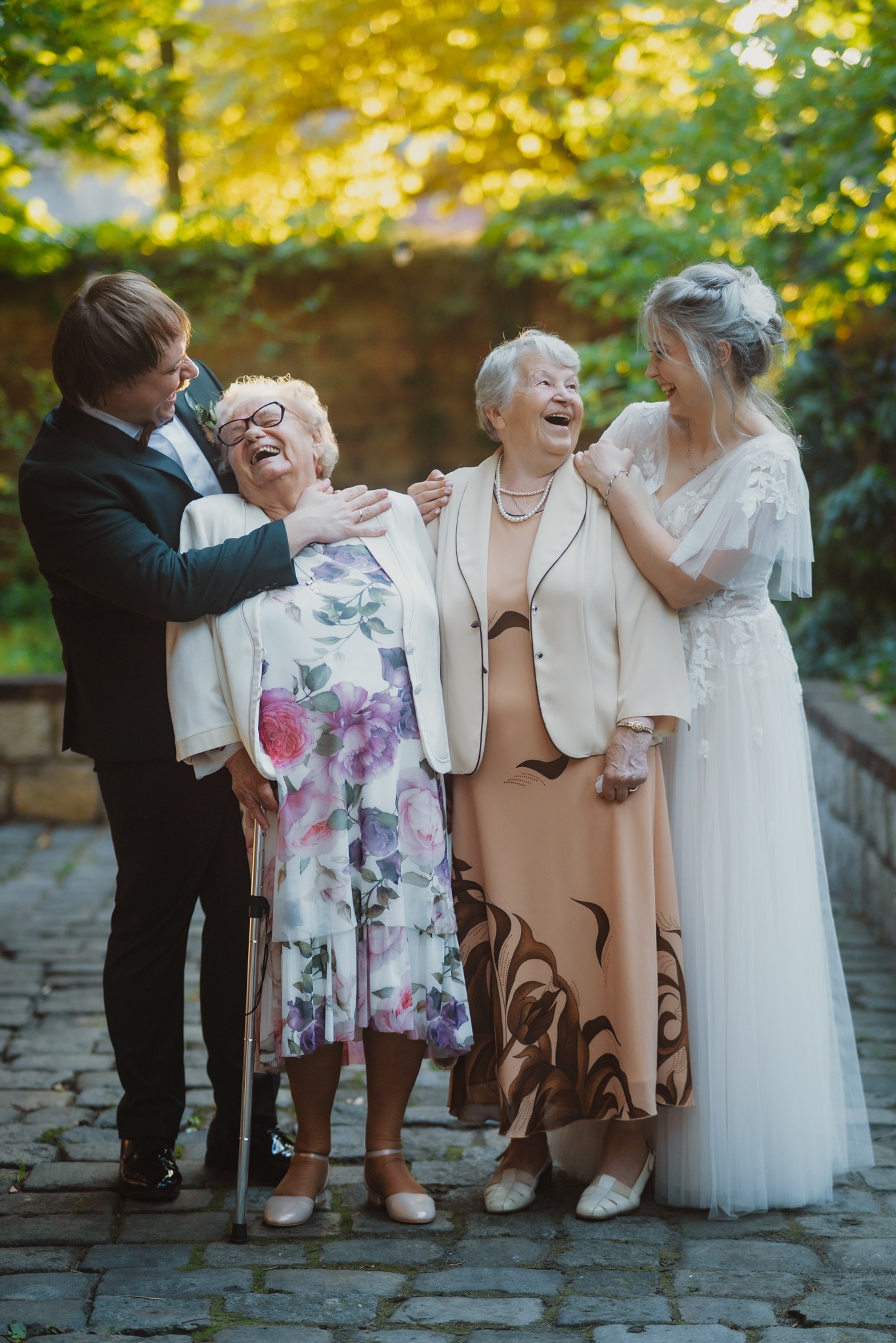 Uśmiechnięta panna młoda w białej sukni i pan młody w garniturze obejmują dwie starsze panie w eleganckich sukienkach na tle zieleni.