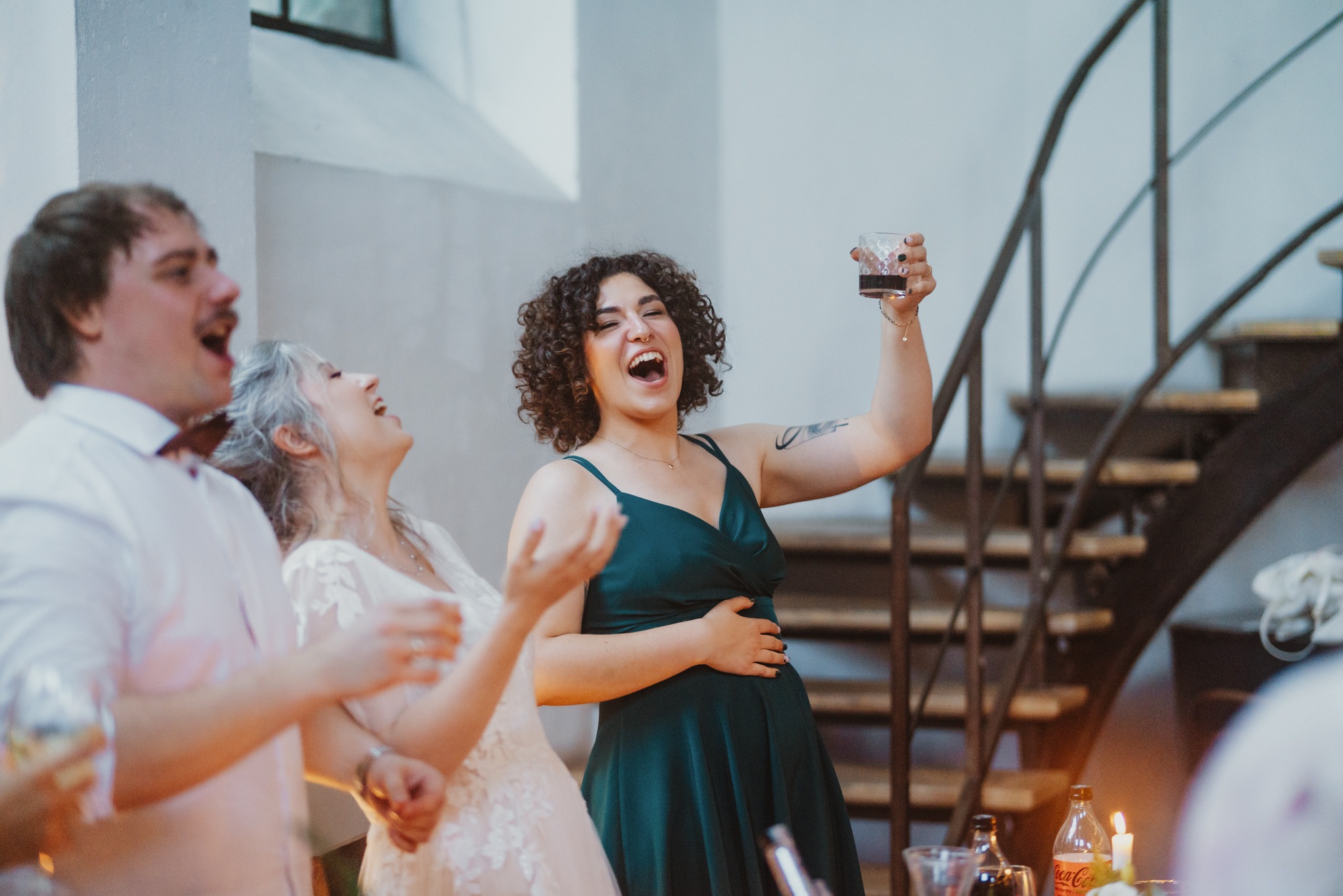 Rozbawiona kobieta w szmaragdowej sukience wznosi toast na tle spiralnych schodów. Obok niej śmieją się goście weselni, tworząc radosną atmosferę.