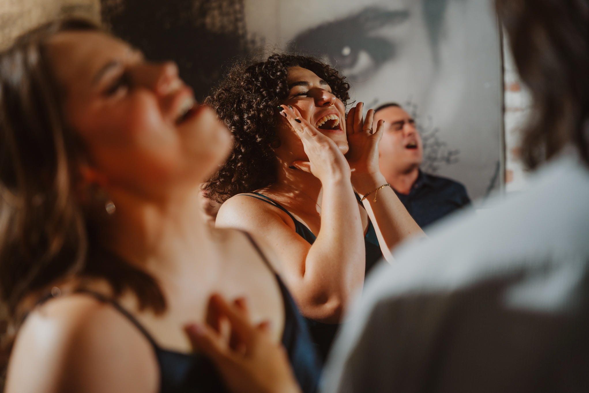 Rozbawiona kobieta z kręconymi włosami śmieje się, trzymając dłonie przy twarzy. W tle widoczne inne osoby, również w radosnych nastrojach. Ujęcie z imprezy.