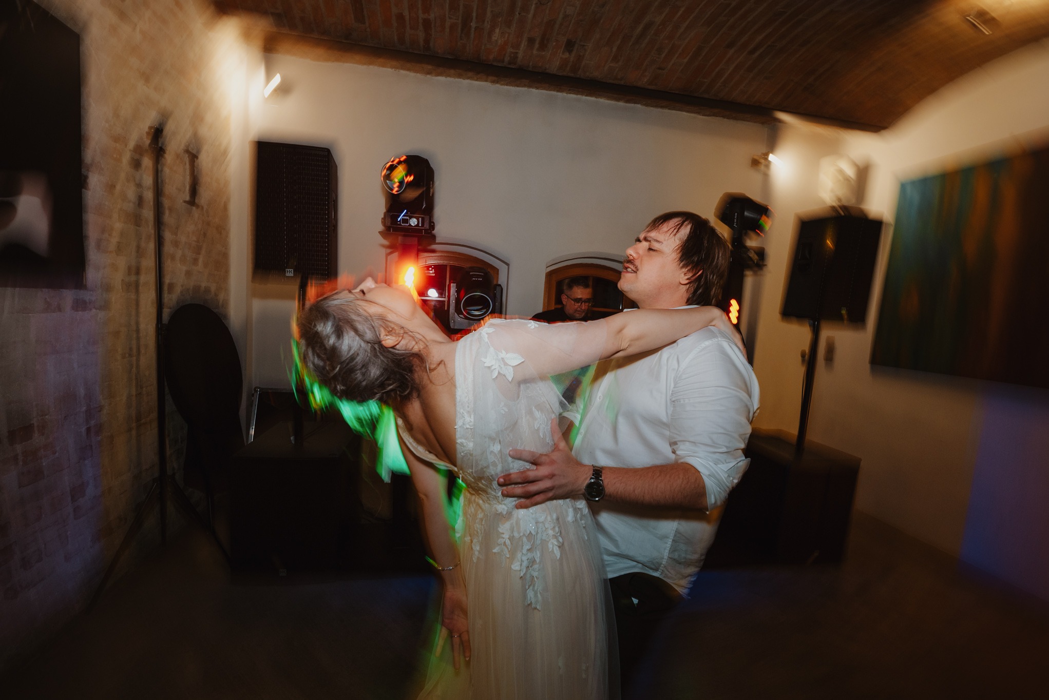 Para w tańcu podczas wesela w Kleszczowie. Panna młoda w białej sukni, pan młody w białej koszuli. W tle DJ i sprzęt nagłośnieniowy, ceglana ściana i efekty świetlne.