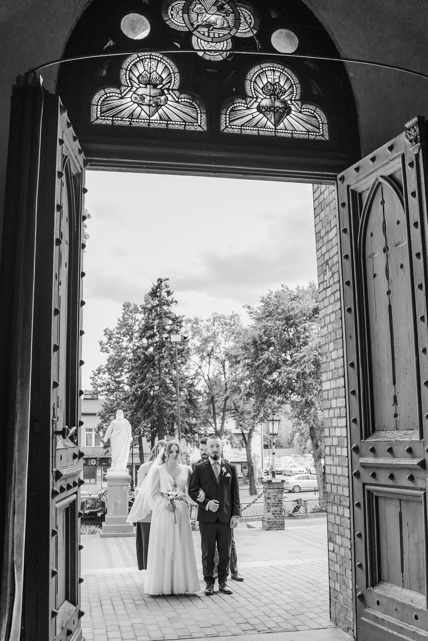 Czarno-białe zdjęcie nowożeńców wychodzących z kościoła przez otwarte, zdobione drzwi. W tle widoczny pomnik i drzewa. Ujęcie z dołu podkreśla architekturę.