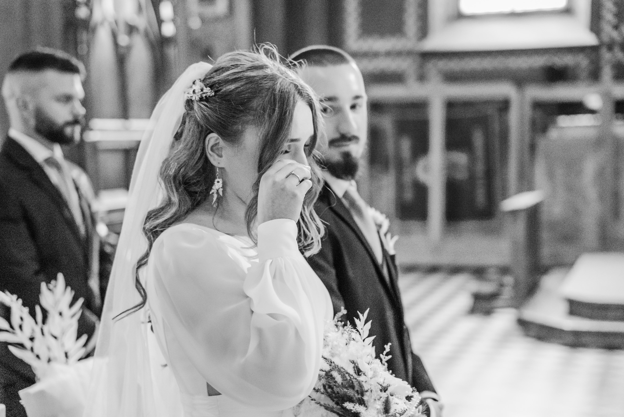 Czarno-białe ujęcie wzruszonej panny młodej ocierającej łzę podczas ceremonii ślubnej w kościele, z widocznym panem młodym i gościem w tle.