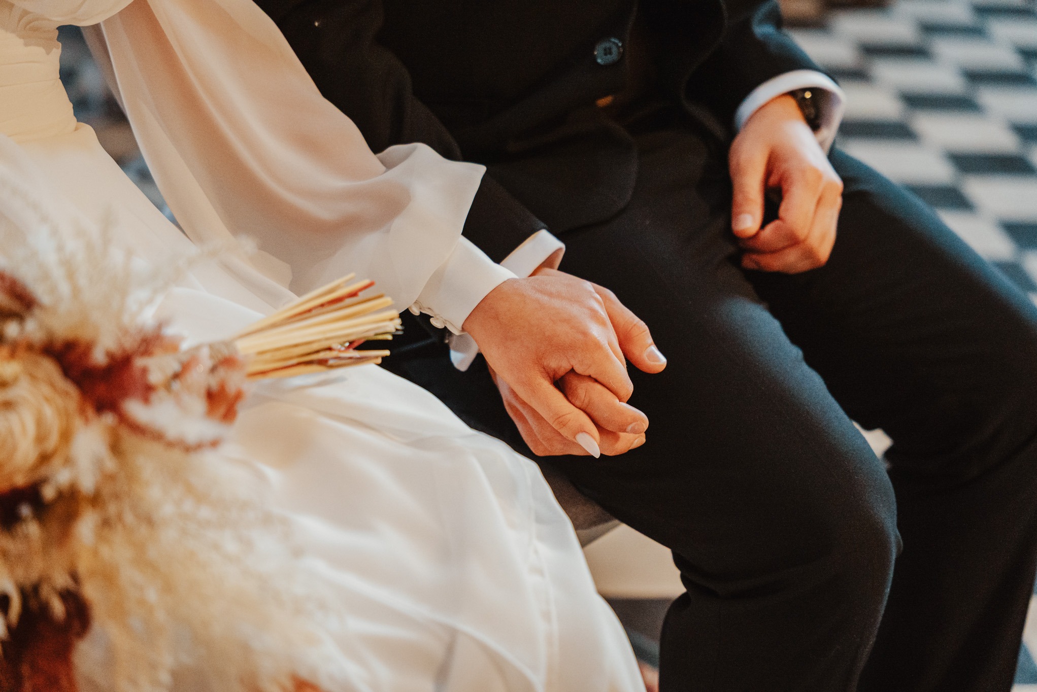 Dłonie pary młodej splecione w intymnym ujęciu, bukiet ślubny z suszonych kwiatów, elegancki strój, subtelne światło podkreśla detale.