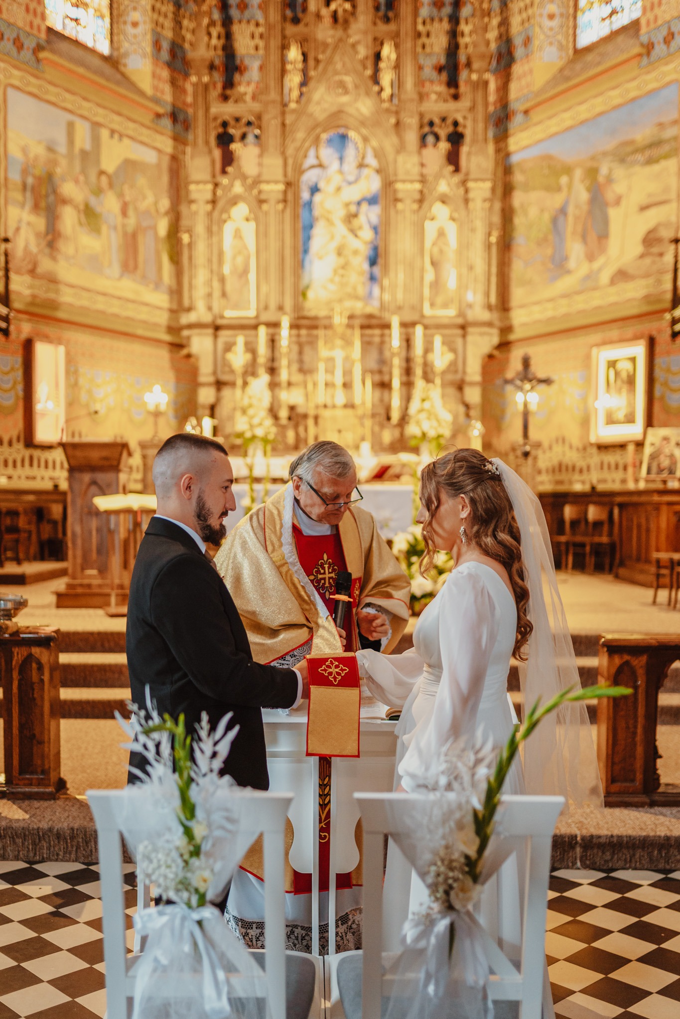 Uroczystość ślubna w kościele: Para młoda podczas ceremonii wymiany obrączek, w tle bogato zdobione wnętrze świątyni i duchowny w szatach liturgicznych.