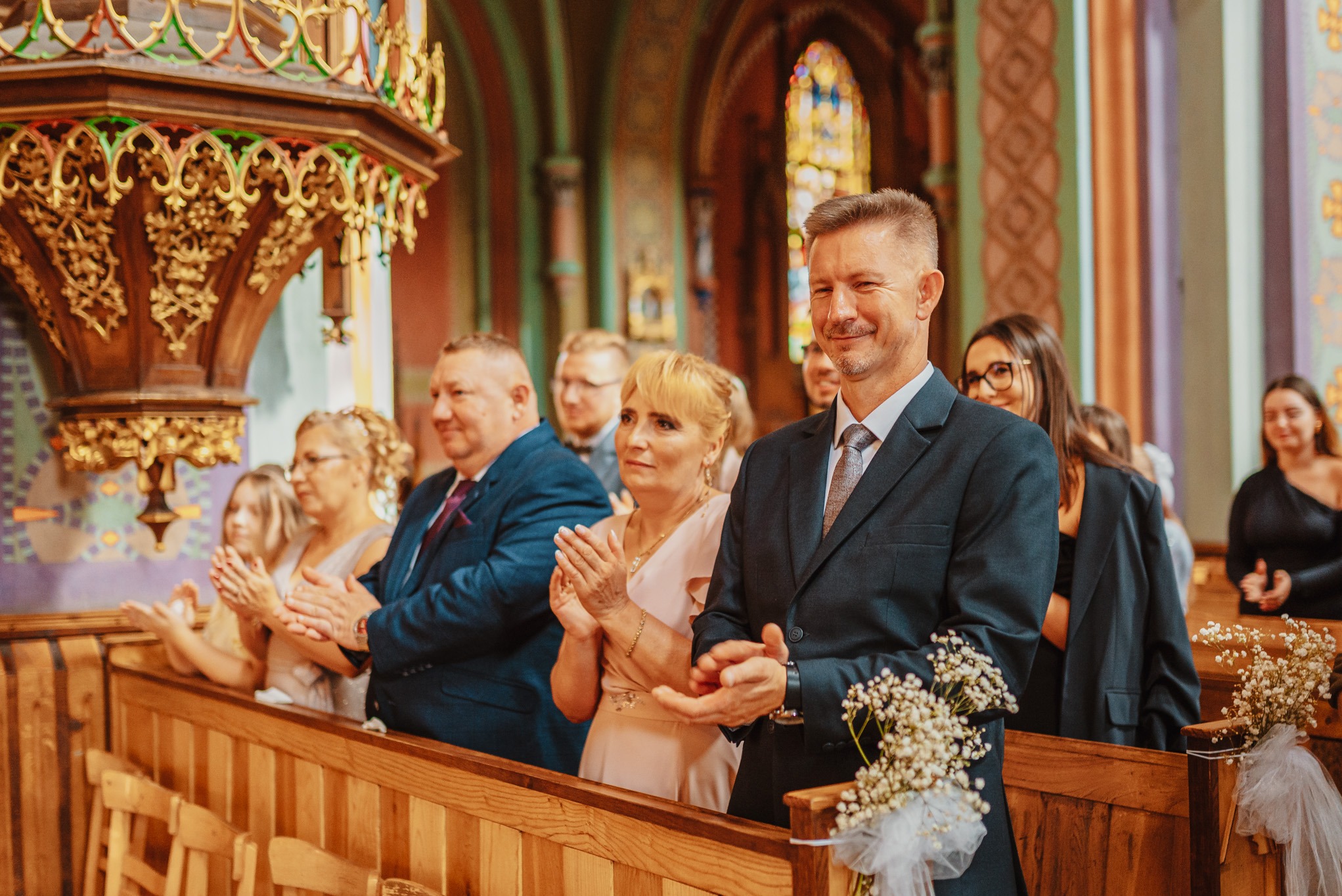 Goście weselni w kościele klasycznym, z drewnianą amboną i witrażami, biją brawo młodej parze. W pierwszym planie mężczyzna w garniturze z bukiecikiem.