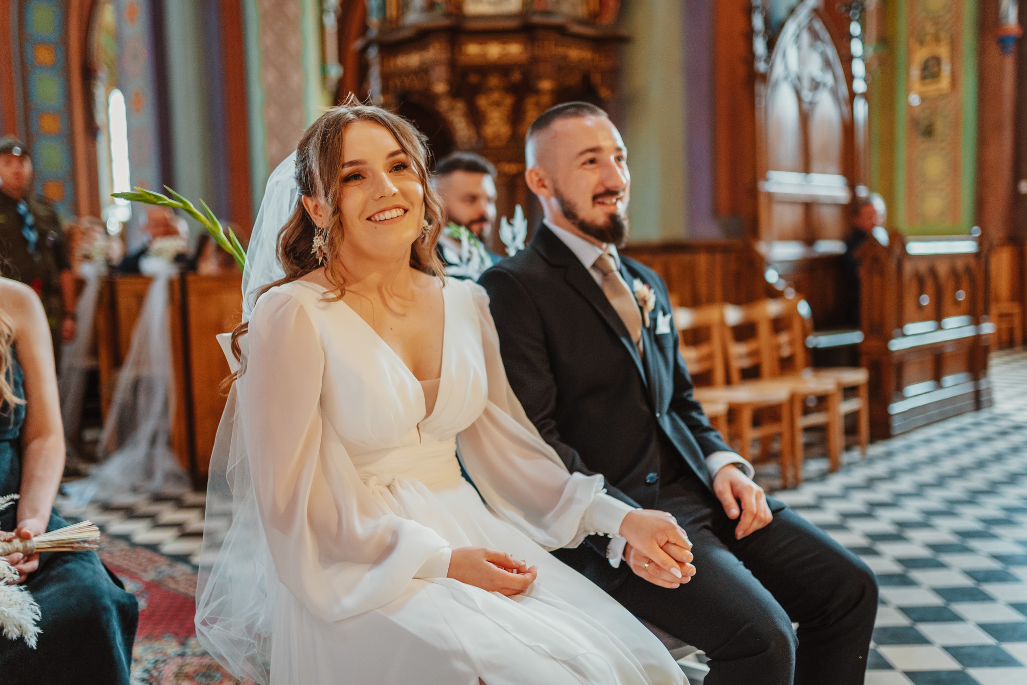 Uśmiechnięta panna młoda w białej sukni i pan młody w czarnym garniturze siedzą obok siebie w kościele, trzymając się za ręce podczas ceremonii ślubnej.