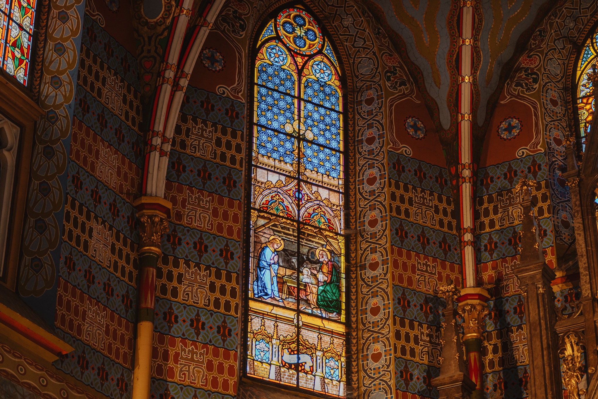 Witraż z motywem religijnym w bogato zdobionym wnętrzu kościoła. Złożone wzory na ścianach i kolumnach, ciepłe oświetlenie podkreśla detale architektoniczne.