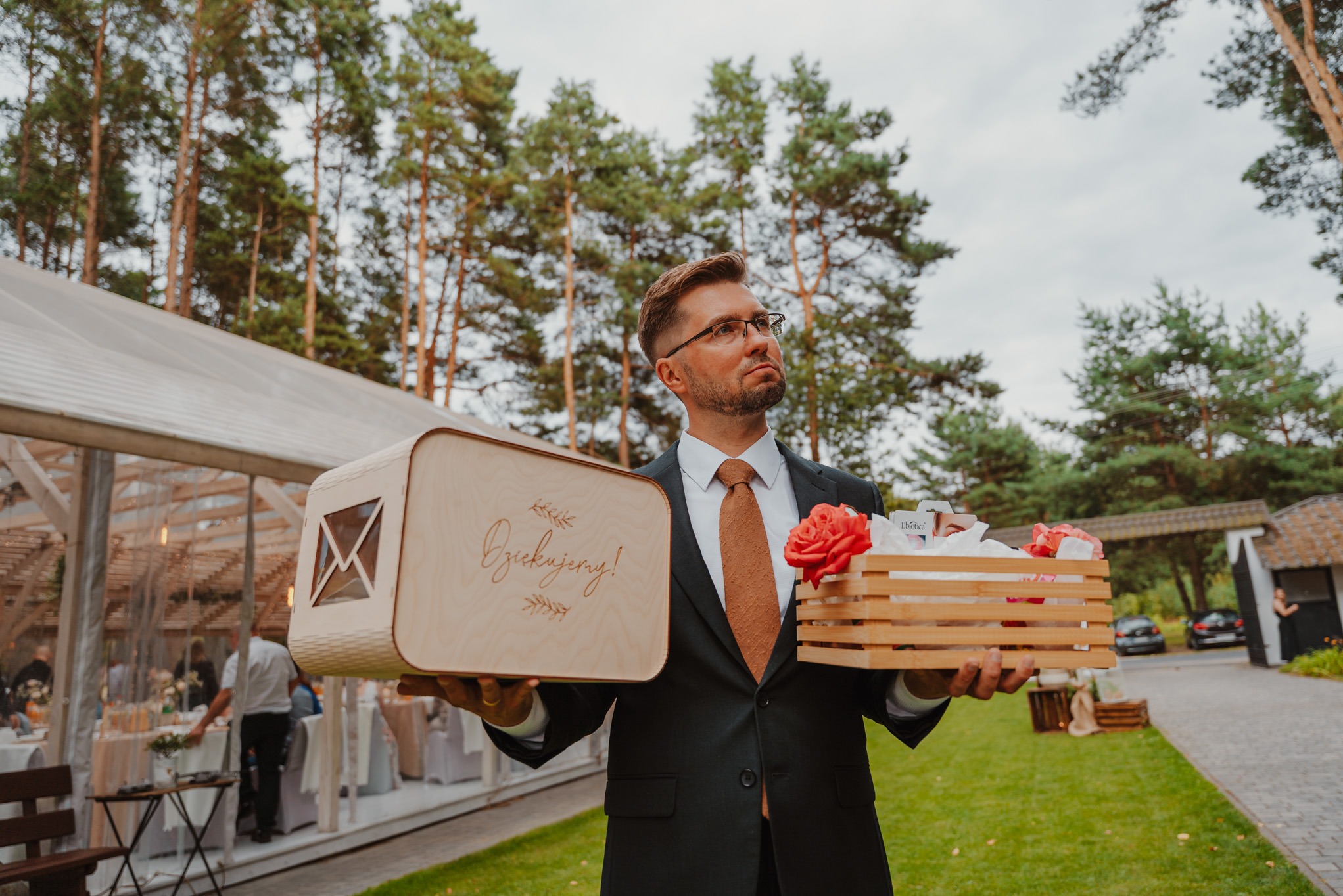 Elegancki mężczyzna w garniturze trzyma drewniane pudełko z napisem 'Dziękujemy!' i skrzynkę z kwiatami na tle plenerowej sali weselnej.