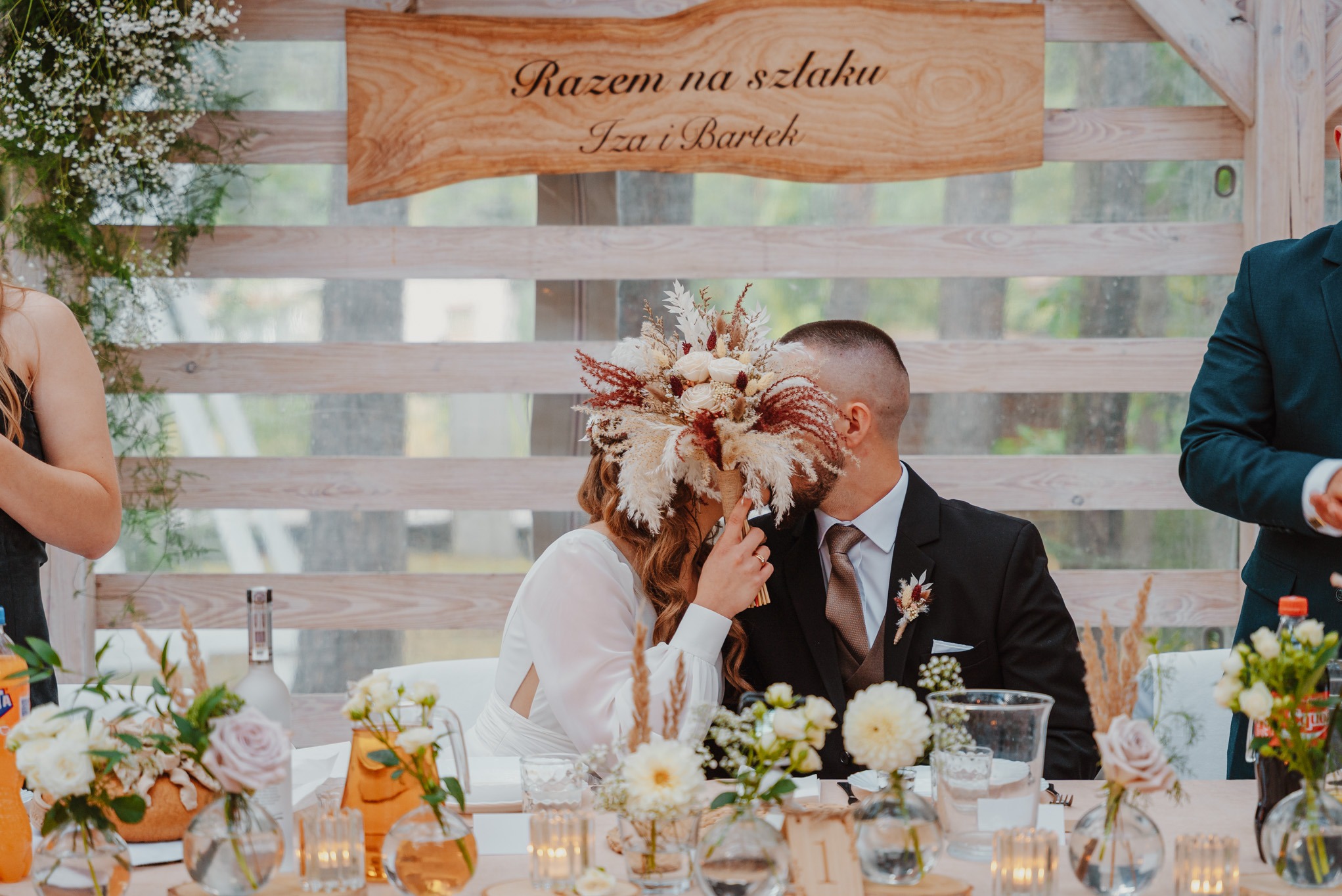 Para młoda w trakcie pocałunku, zasłonięta bukietem z suszonych kwiatów, siedząca za stołem z dekoracjami kwiatowymi i świecami, w tle drewniana tablica z napisem.
