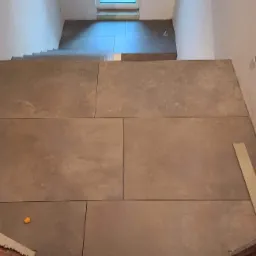 Widok z góry na schody i podest wyłożone dużymi, szarymi płytami. Widoczne niewielkie okno na końcu schodów i fragment listwy przypodłogowej.