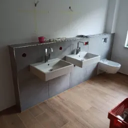 Dwie białe, kwadratowe umywalki z chromowanymi kranami, zamontowane na szarych płytkach ściennych, z bidetem po prawej stronie; na ścianie widoczne ślady po taśmie malarskiej, a na blacie poziomica...