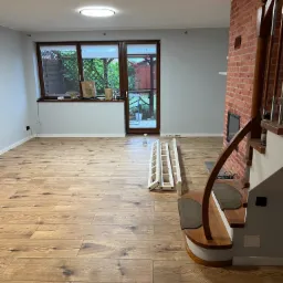 Wnętrze pokoju z nową drewnianą podłogą, widoczne schody z drewnianą balustradą, ceglany kominek, okno z widokiem na ogród i rozłożona drabina malarska.