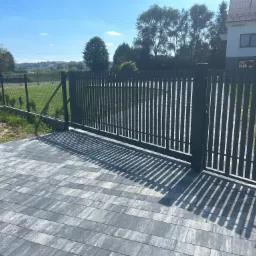 Nowoczesna, grafitowa brama wjazdowa z pionowymi sztachetami, otwierana automatycznie, z widoczną kostką brukową na podjeździe i domem w tle.