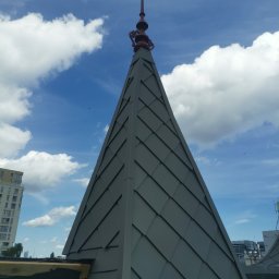 TAKI DACH - Szpiczasty dach z blachy w Olsztynie, zakończony ozdobnym elementem, na tle błękitnego nieba z chmurami.