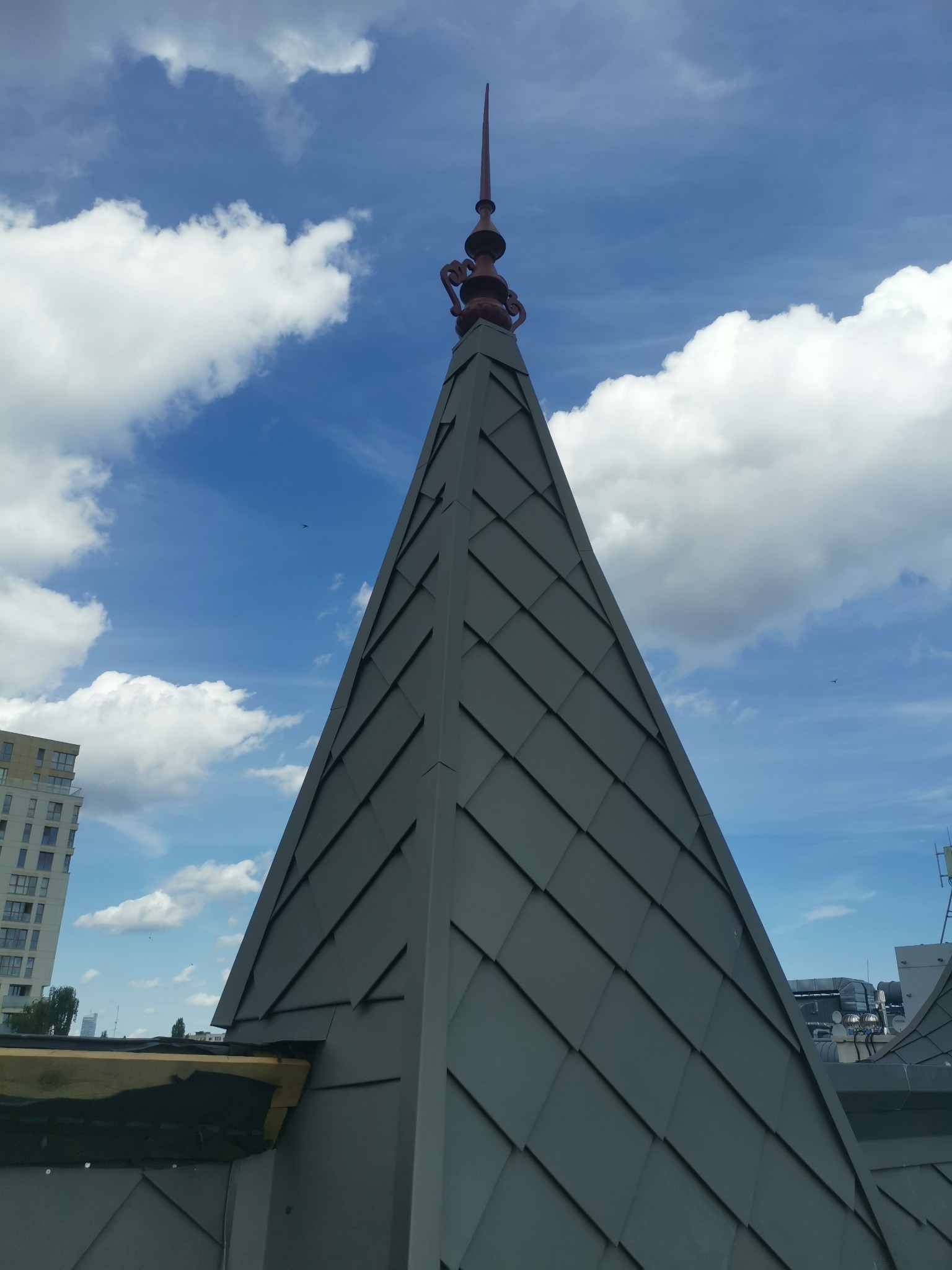 Szpiczasty dach z blachy w Olsztynie, zakończony ozdobnym elementem, na tle błękitnego nieba z chmurami.