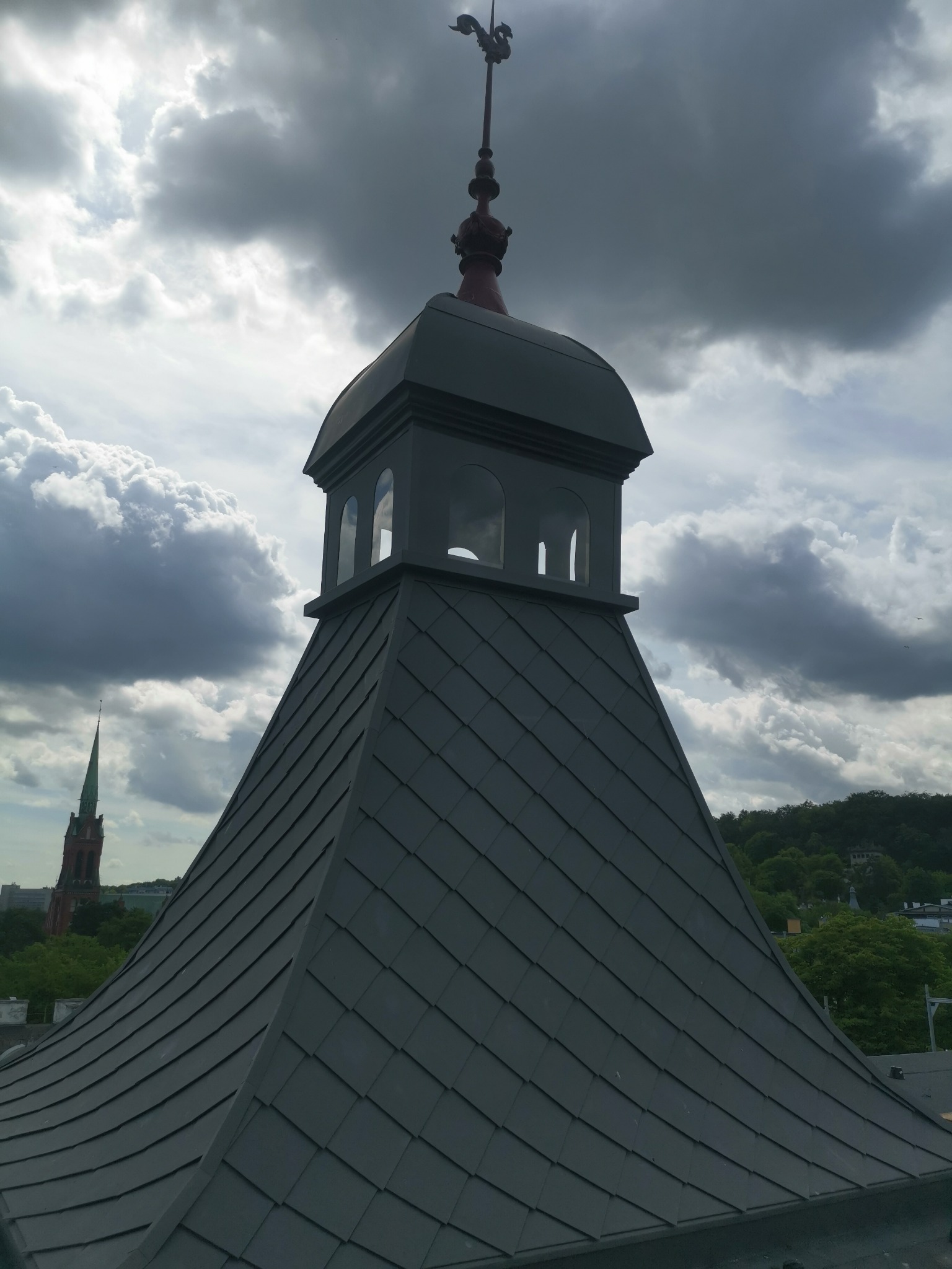 Szary dach wieżyczki z metalowymi płytkami, zwieńczony czerwonym elementem dekoracyjnym z wiatrowskazem, na tle burzowego nieba i odległej wieży kościelnej.