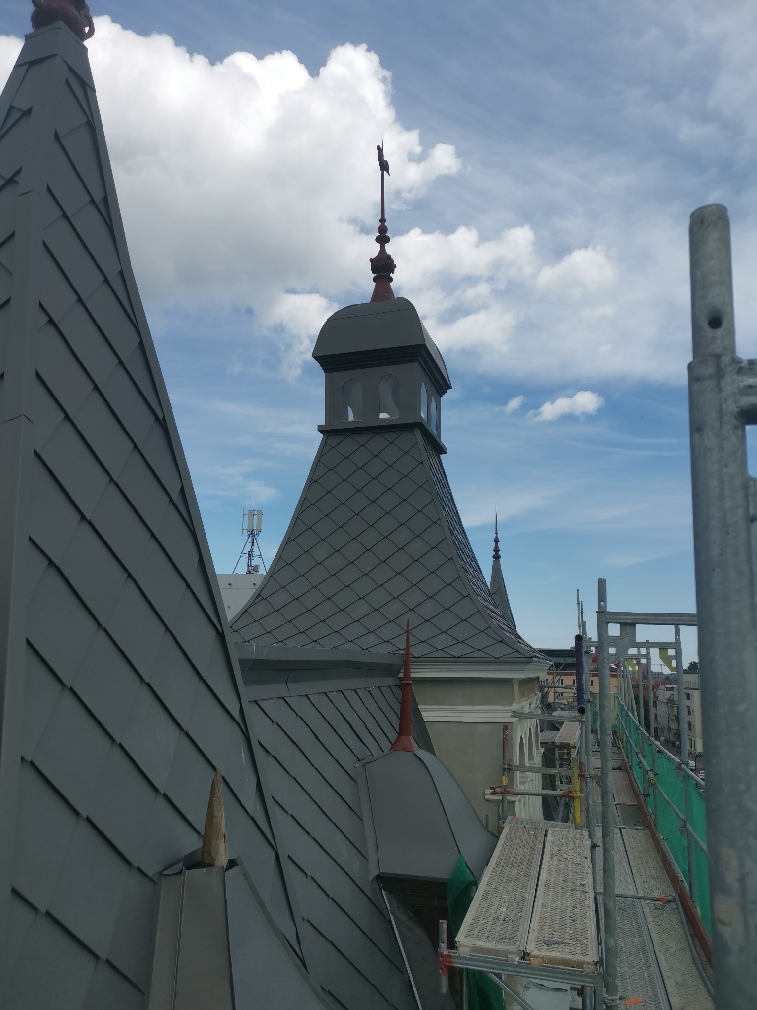 Szare dachy z blachy trapezowej i wieżyczka z iglicą, widoczne rusztowania i fragmenty miasta w tle, pochmurne niebo.
