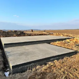 Dwa betonowe fundamenty w wykopie, zabezpieczone czarną izolacją, na tle zaoranego pola i błękitnego nieba z pojedynczym pracownikiem w oddali.