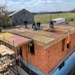 Wznoszenie ścian domu z czerwonej cegły ceramicznej z widocznym zbrojeniem stropu i pracownikami na górze, w tle budynek gospodarczy i zielone pola.
