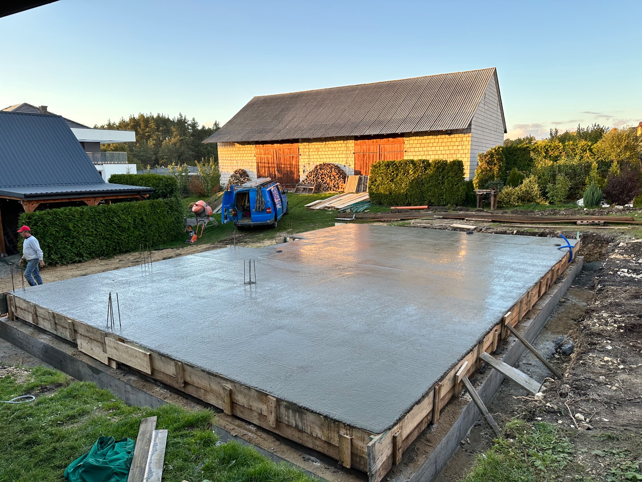 Wylany, świeży beton na przygotowanym fundamencie z drewnianym szalunkiem, widoczne wystające pręty zbrojeniowe, w tle budynek gospodarczy z drewnem opałowym i zaparkowany niebieski bus.